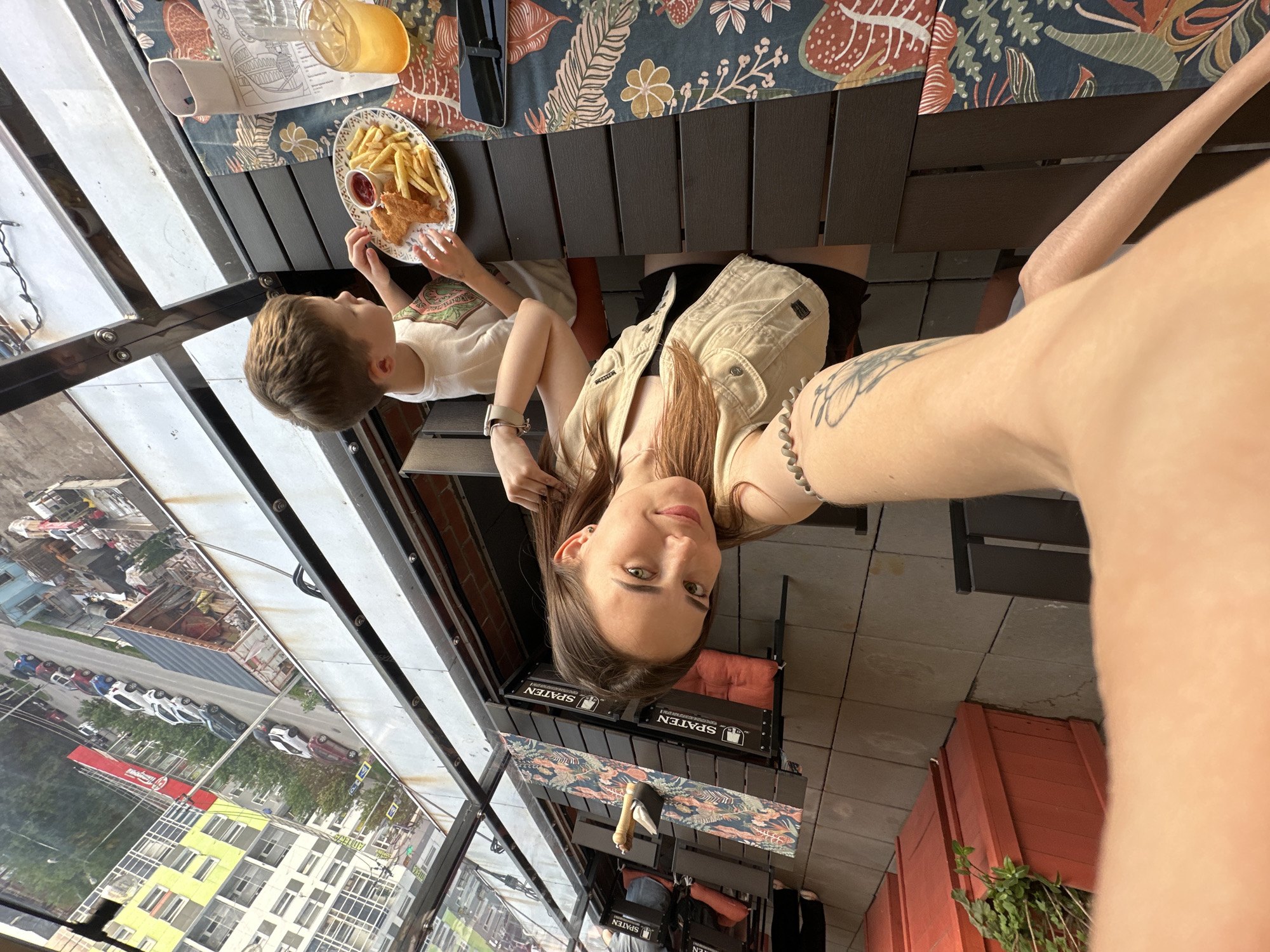 A woman is taking a selfie while at a restaurant in Perm, Russia. The restaurant has an outdoor seating area with a view of the city. The woman is wearing a beige jacket and has brown hair. A young boy, presumably her son, is sitting across from her and eating french fries and fried chicken. The table has a patterned tablecloth and there is a glass of orange juice on the table. The city of Perm is visible through the glass enclosure of the restaurant, showing buildings, cars, and trees.
