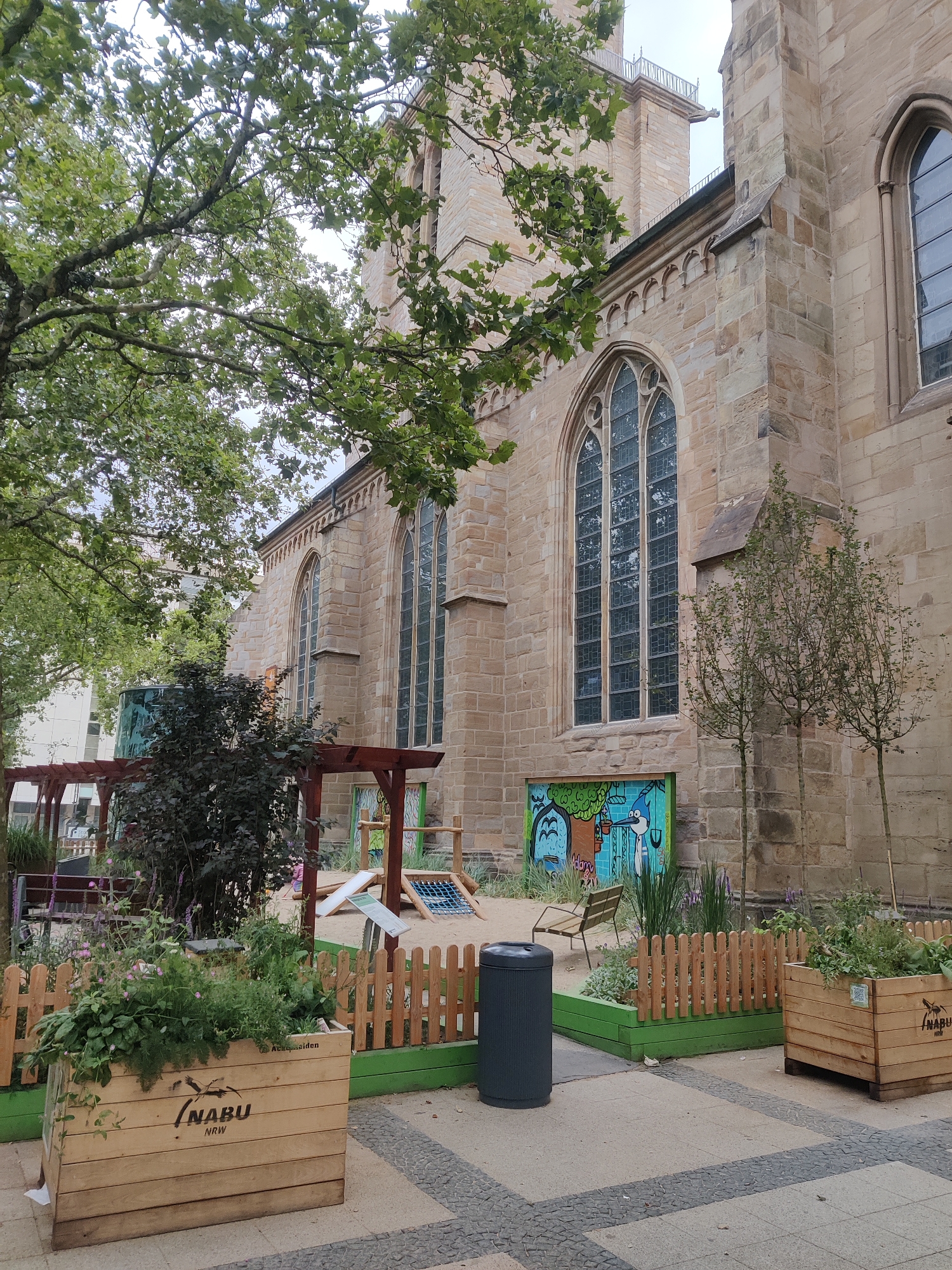The image depicts the exterior of St. Reinoldi's Church in Dortmund, Germany. The stone facade of the church, characterized by large Gothic-style windows and intricate stonework, is visible. In the foreground, a vibrant urban garden area is set up. This garden features wooden planters filled with lush green plants, a small wooden fence, a black trash bin, and a playground structure. A colorful mural adorns the lower part of the church wall, adding a touch of art to the scene. Trees with green foliage are scattered throughout the area, providing shade and framing the view of the church. The ground is paved with stone tiles. No people are visible in the image.