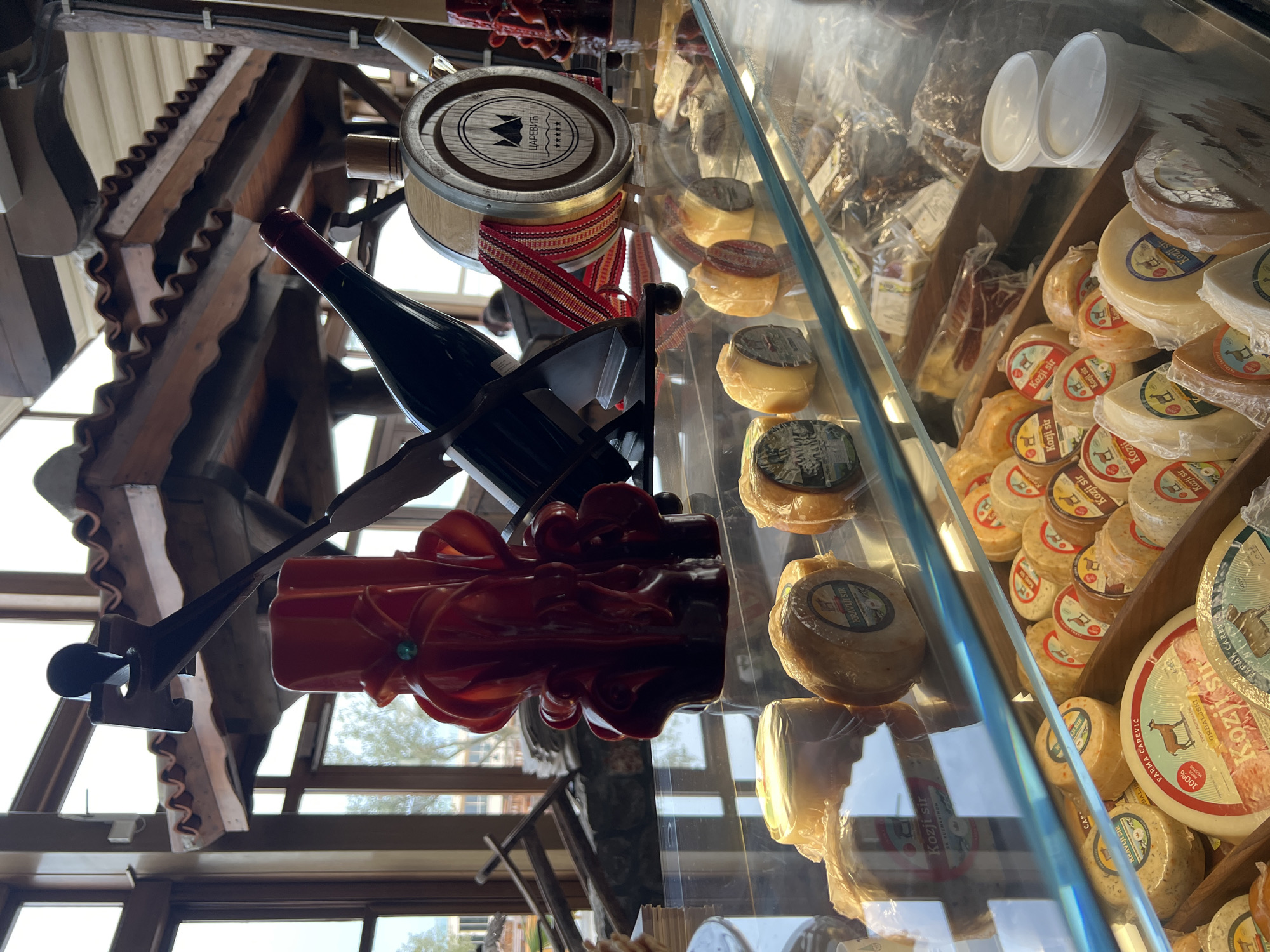 This image showcases a display of local products, likely within a store or market in Kovačko Polje, Montenegro. The foreground features a glass display case filled with an assortment of cheeses, many of which appear to be goat cheeses ("Kozji Sir"). The packaging and labels on the cheeses are in the local language. Above and behind the display, there is a decorative arrangement involving a bottle of dark red wine, a wooden barrel with local branding ("ЦАРЕВИЋ"), and a large, intricately sculpted red candle, all held together by a metal stand. The location is indoors with the structure of a wooden roof visible. Outside the windows can be seen what appears to be a building across the street.