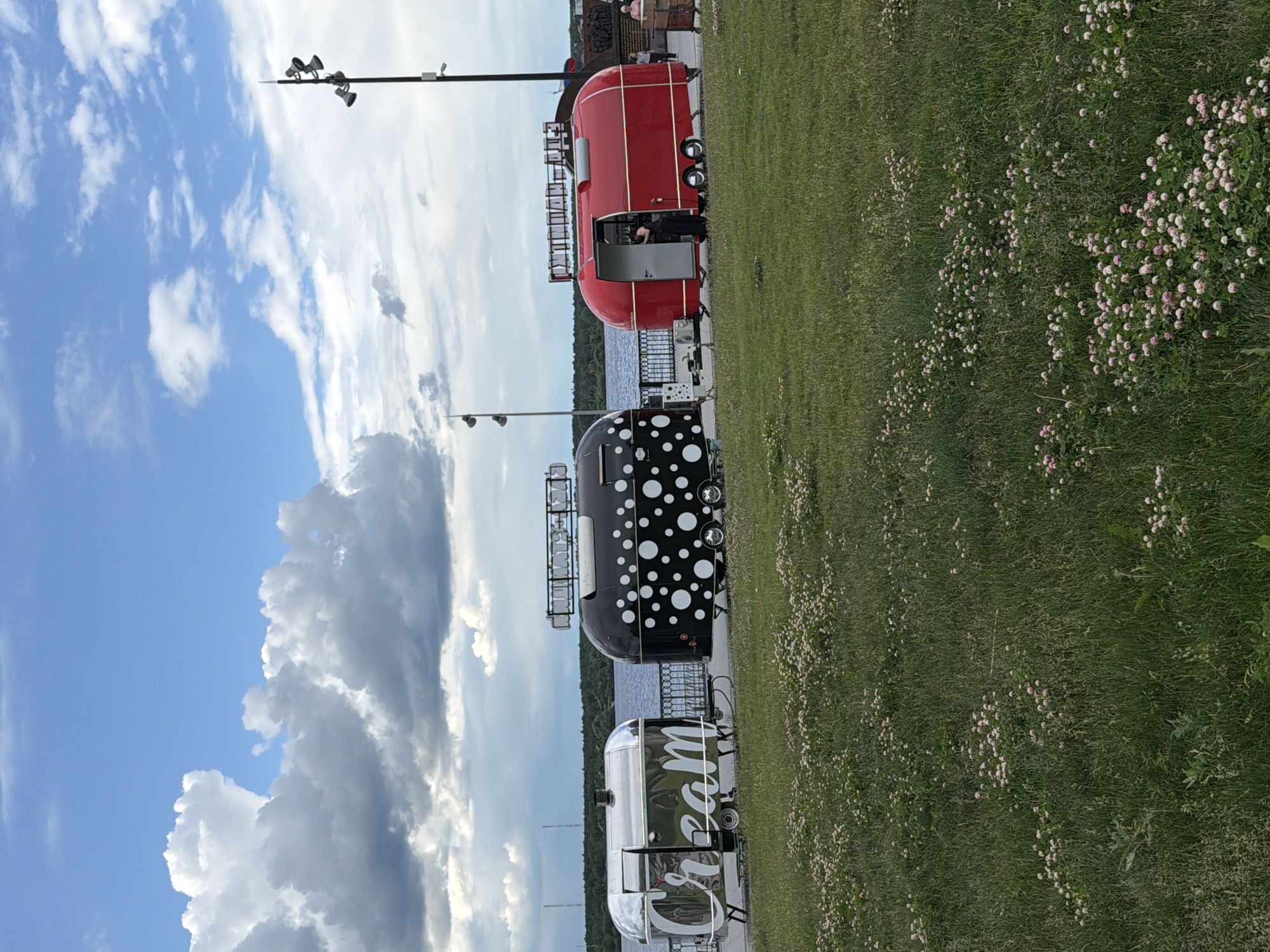 The media file displays a scene with three food trailers lined up along a grassy area next to a body of water. The sky is a mix of blue with white clouds. The trailers have distinct designs: one is bright red, another is black with white polka dots, and the third is metallic and silver with "Cream" written on its side in large white lettering.
In the red trailer, a person is visible inside, seemingly working. The trailers appear to be set up for business, suggesting they are part of a food stall or event. The grassy area is dotted with small pink and white flowers, indicating it's likely springtime or summer. The presence of a lake or river suggests an outdoor recreational area. The lighting implies it is daytime, possibly late afternoon or early evening, given the angle of the light and cloud formations. The overall impression is a pleasant, sunny day in an outdoor setting, likely a public space in Perm, Russia, where food vendors are operating. There is no overt indication of a specific event, but the setup suggests a commercial or community gathering.