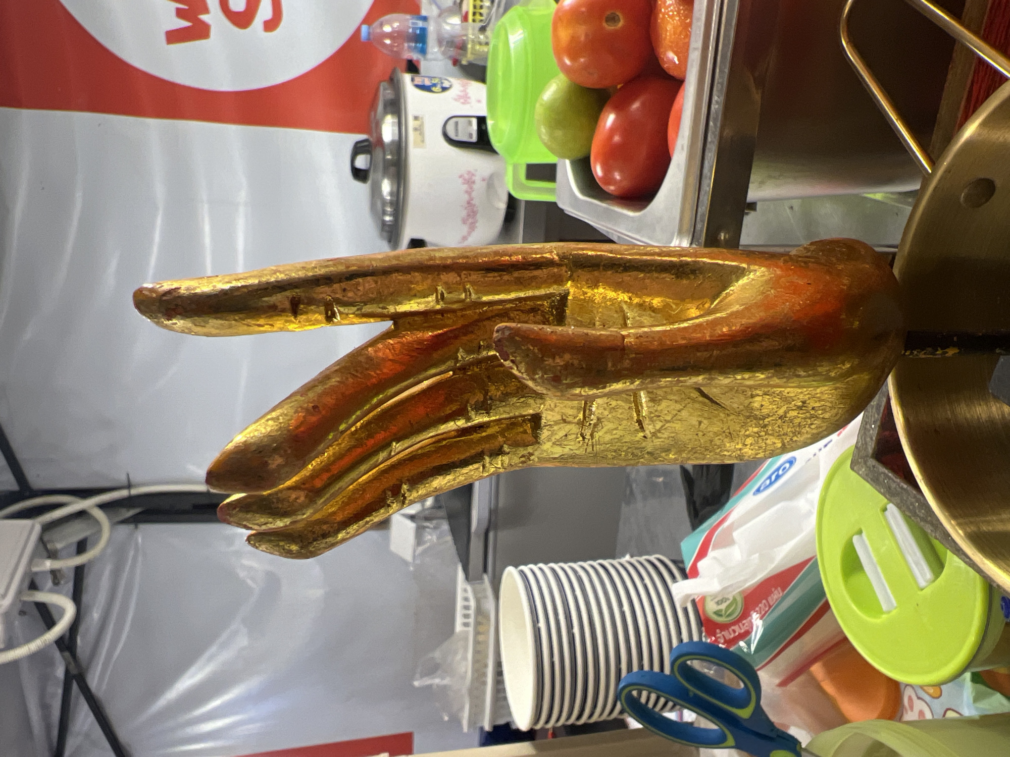 The image shows a close-up, rotated view of a golden hand sculpture in the foreground. The sculpture appears to be made of metal with a textured, reflective surface. The hand is in a gesture with the index finger extended. In the background, there are various objects on a shelf or counter. These include a white rice cooker with a metal lid, a clear plastic bottle of water, some red tomatoes and a green fruit in a metal container, and a stack of black and white striped paper cups. A green plastic lid with white indentations is also visible, along with a pair of blue scissors with red accents. Several plastic bags and wrappers are scattered around, one of which has visible text. The overall setting appears to be a casual commercial or domestic space, possibly a stall or a kitchen area, given the presence of cooking appliances and food items. The lighting suggests it could be daytime, but there are no clear indications of weather or a specific time. The location context is Karon, Thailand.