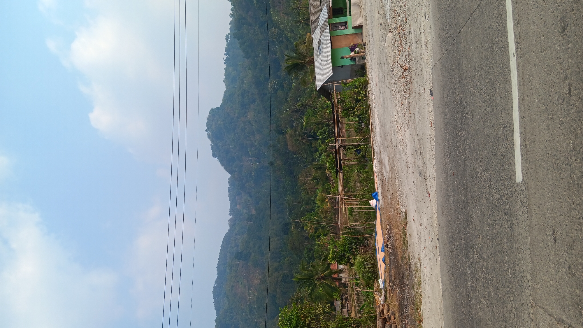 The image shows a rural scene in Mekargalih, Indonesia, with a paved road in the foreground and a lush green mountainside in the background. A few simple houses, some with corrugated metal roofs, are visible among the trees. Electrical wires stretch across the frame above the landscape. In the middle ground, there are areas of cleared land with what appear to be building materials and tarps, suggesting ongoing construction or renovation. A person is standing near one of the houses. The sky is partly cloudy with a light blue hue.