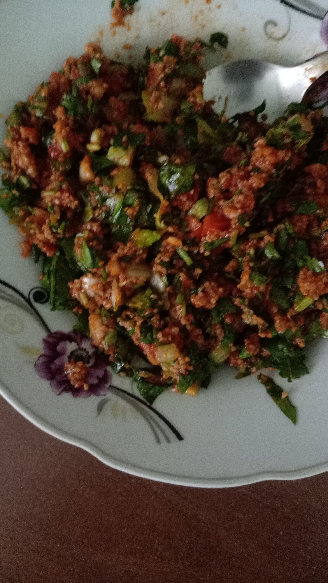 A close-up shot shows a plate of food, possibly Çiğ Köfte, on a table. The plate is decorated with a purple flower and has curved lines. The food consists of small, reddish-brown grains mixed with chopped green vegetables. A silver spoon is partially visible in the upper right corner of the plate. The table is a solid brown color. The location is likely Yerevan, Armenia.