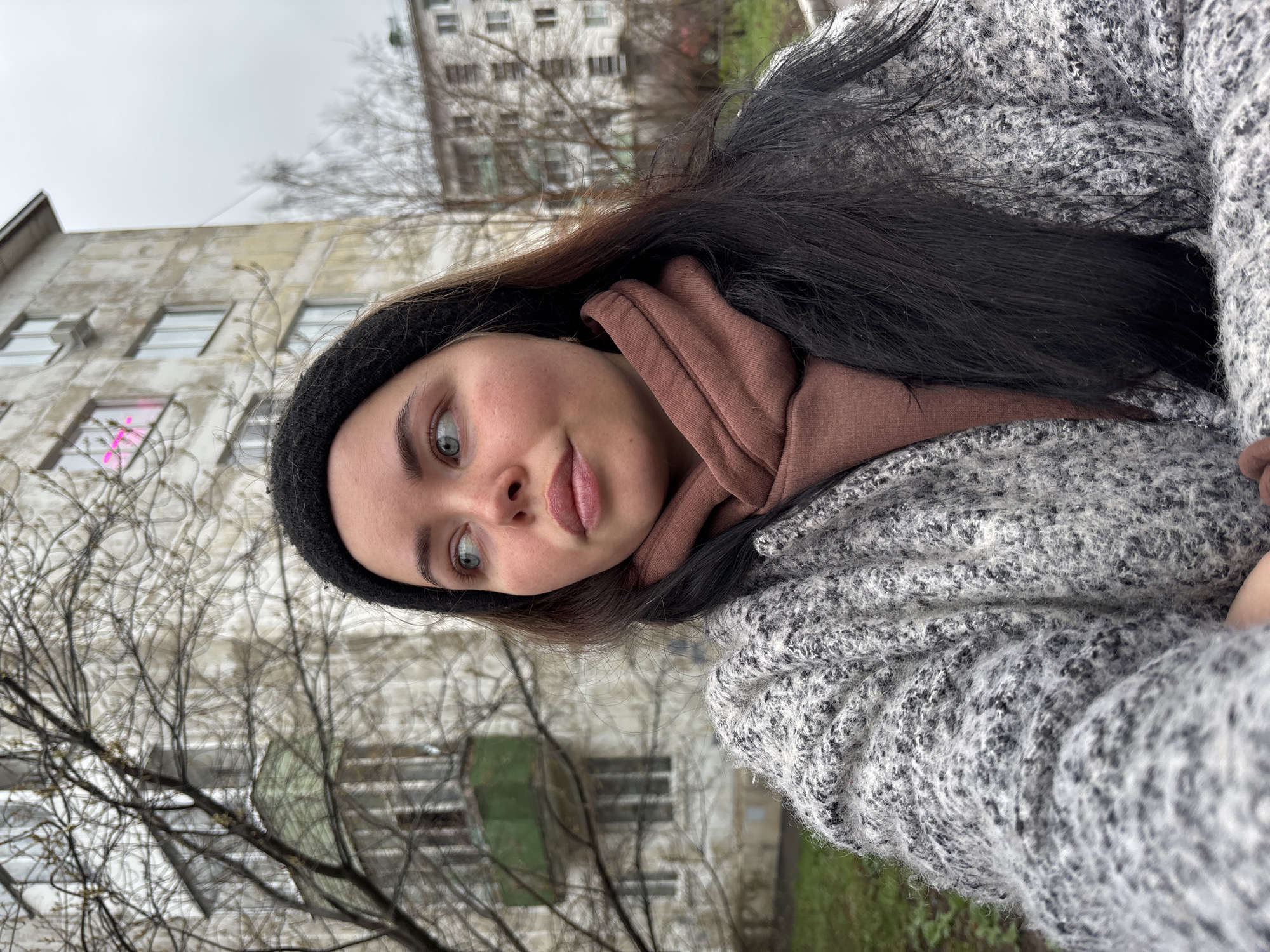 The image is a portrait of a young woman, rotated 90 degrees counter-clockwise, capturing her from the chest up in an outdoor urban setting. She has long, dark hair, light greenish-blue eyes, and a fair complexion, looking directly towards the camera with a subtle, slightly contemplative expression. Her lips are slightly parted.
She is dressed for cool weather, wearing a dark knit beanie, a brown hooded sweatshirt, and a thick, speckled light gray and black knitted sweater or cardigan. Her hair falls over her shoulder and is partially covered by the beanie and sweater.
The background features several multi-story buildings, typical of utilitarian residential or administrative architecture, with visible windows. One building shows an air conditioning unit below a window, and another has a section of its facade colored dark green. On a window of one of the buildings, faint pink graffiti-like marks are visible. Bare tree branches are prominent in the foreground and midground, indicating a season like late autumn, winter, or early spring. The sky is overcast and uniformly gray, suggesting a cloudy day.
The scene conveys a casual, everyday moment in a city environment, consistent with the provided location context of Izhevsk, Russia. The overall color palette is muted, dominated by grays, browns, and the natural tones of the urban landscape.