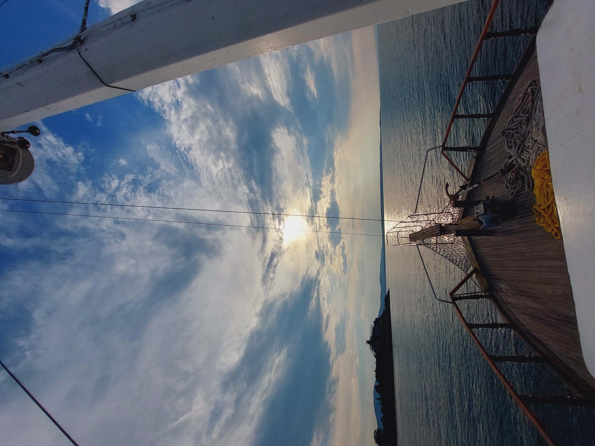 The image depicts a view from the bow of a wooden ship, sailing on a calm sea towards a distant island. The sun is partially obscured by clouds, casting a bright glare on the water. The sky is a mix of blue and white clouds, with sunlight filtering through. On the left side of the image, a silhouette of the island with a building is visible against the horizon. The deck of the ship is made of wood, with railings and ropes visible. The scene appears to be the coast of Sozopol, Bulgaria, specifically the area around the Old Town.