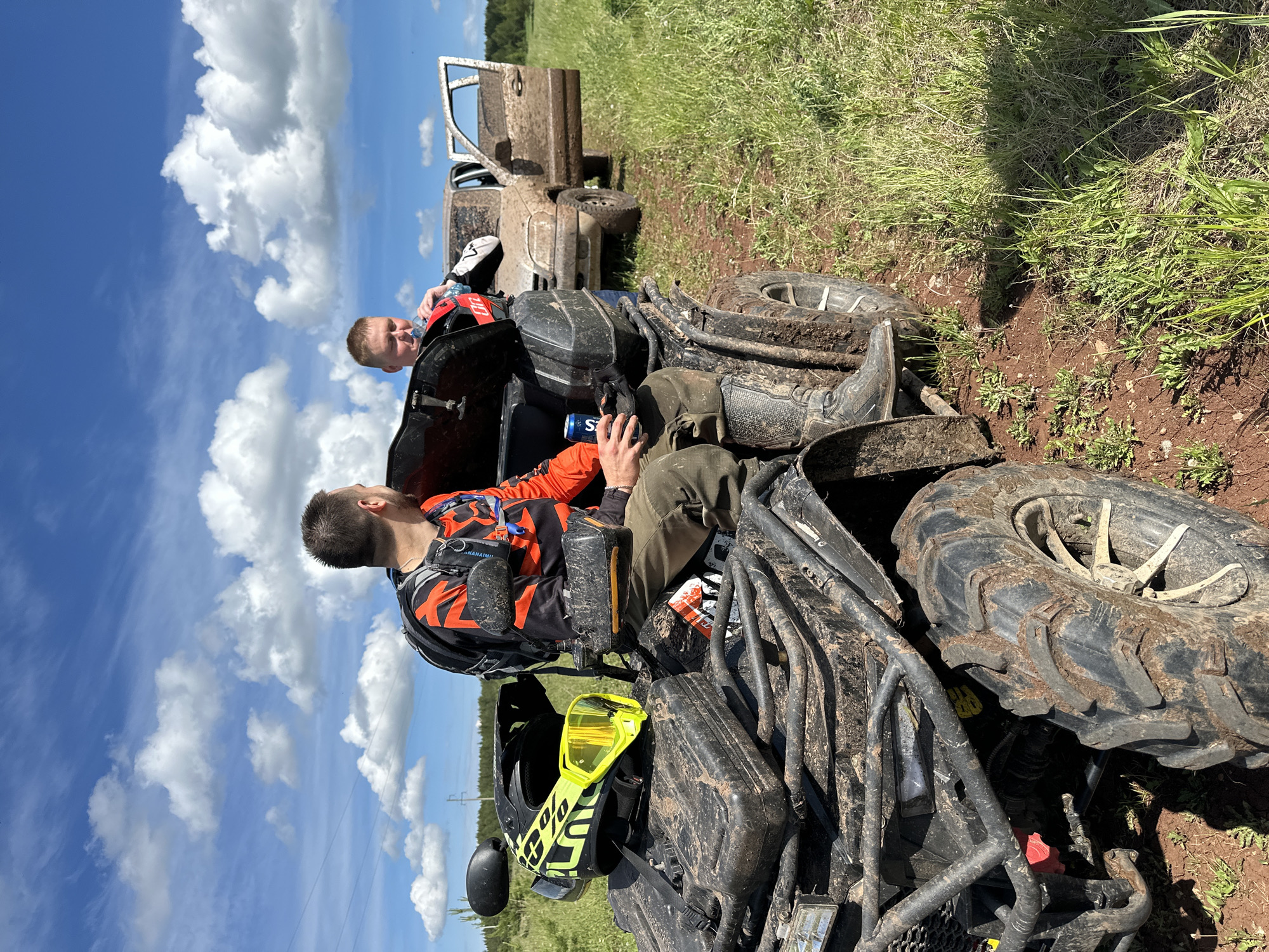 Two individuals are near a muddy ATV on a grassy field, with a dirty white vehicle in the background, under a partly cloudy sky. The location is not identifiable as Ilyinsky, Russia from this image. One person, wearing an orange and black shirt, is sitting on the ATV, holding an energy drink can. The other person, wearing a black and white jacket, is seated on the ATV's rear. A black helmet with yellow goggles rests on the front of the ATV. The environment is predominantly green with evidence of muddy terrain.
