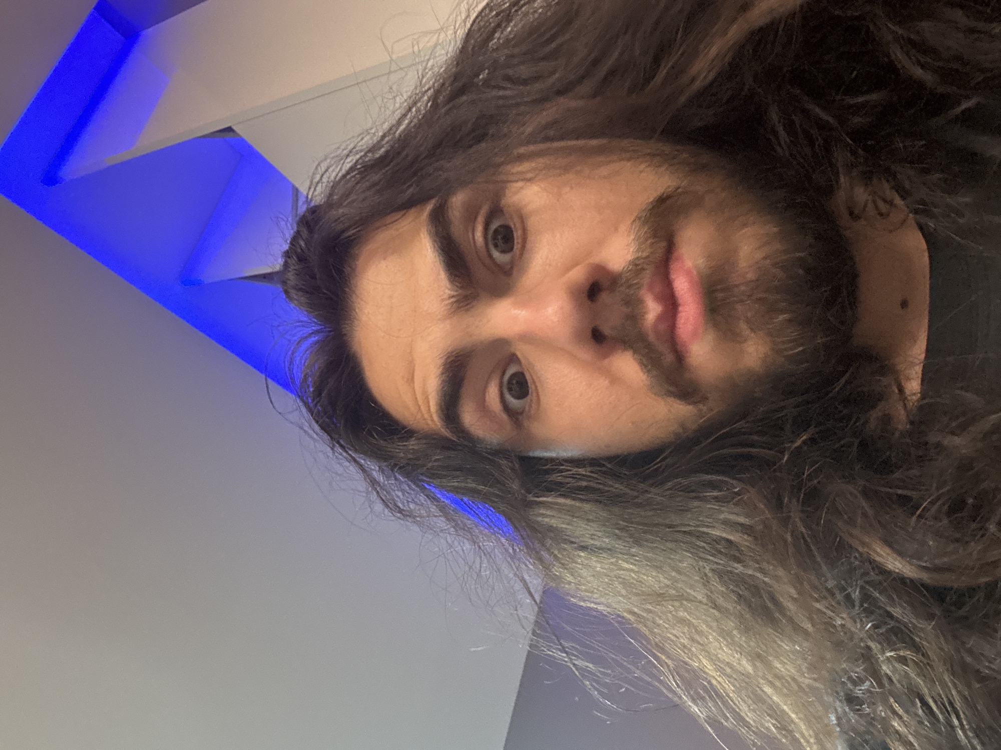 A close-up, high-angle, indoor, selfie-style photograph shows a person with long, dark, messy hair and a beard. The person's eyes are wide open, and their mouth is slightly ajar. The background features a ceiling with blue lighting and a white architectural element. The photograph appears to be taken in the city of Unknown, Unknown.