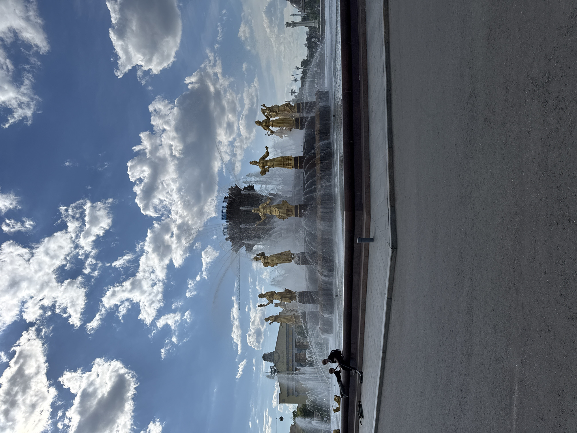A grand, multi-tiered fountain, adorned with numerous golden statues, dominates the central and left portions of the image. The statues depict female figures in various poses, some holding sheaves of wheat or other symbolic items, and are arranged around a central column within the fountain's basin. Water actively sprays from multiple jets, creating a vibrant display of mist and flowing water around the intricate golden sculptures.
Above the fountain, a clear, bright blue sky is visible, scattered with large, fluffy white cumulus clouds. In the distant background, behind the fountain, faint architectural elements of a large building or pavilion can be seen, characterized by light-colored facades and a prominent dome.
The right side and foreground of the image feature a wide expanse of dark, textured pavement, likely asphalt, suggesting a broad walkway or plaza alongside the fountain. In the lower left foreground, a few dark figures are visible; one appears to be a person operating a camera or video equipment on a tripod, indicating someone is filming or photographing the scene.
The scene is bathed in bright daylight, suggesting a sunny day, likely midday or early afternoon. This iconic fountain, with its distinctive golden figures and surrounding architecture, is identifiable as the Friendship of Peoples Fountain at VDNKh in Moscow, Russia. No visible text is discernible in the image.