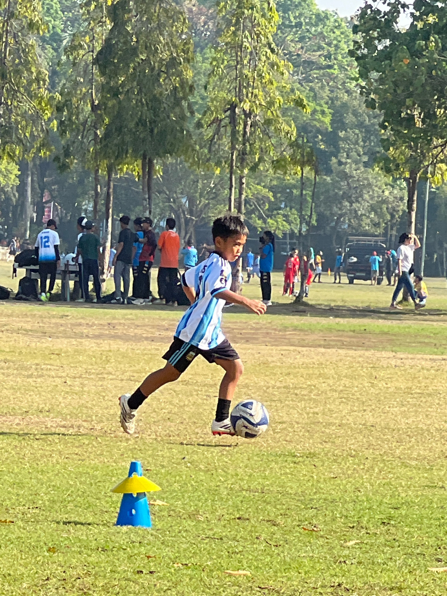 A young boy in a blue and white striped soccer jersey and black shorts dribbles a soccer ball on a grassy field. He is in motion, with his right leg extended forward and his left leg bent. His body is angled to the right, suggesting he is running or moving with the ball. The soccer ball is to his right, in contact with his left foot. In the foreground, a blue and yellow training cone sits on the grass. In the background, several other people are visible, some standing and watching, others appearing to be involved in activities. Trees and foliage form a backdrop, with a dark-colored vehicle and a picnic table also present. The lighting suggests it is daytime, likely sunny. The context of Denpasar, Indonesia, suggests this scene takes place outdoors in a park or sports facility.