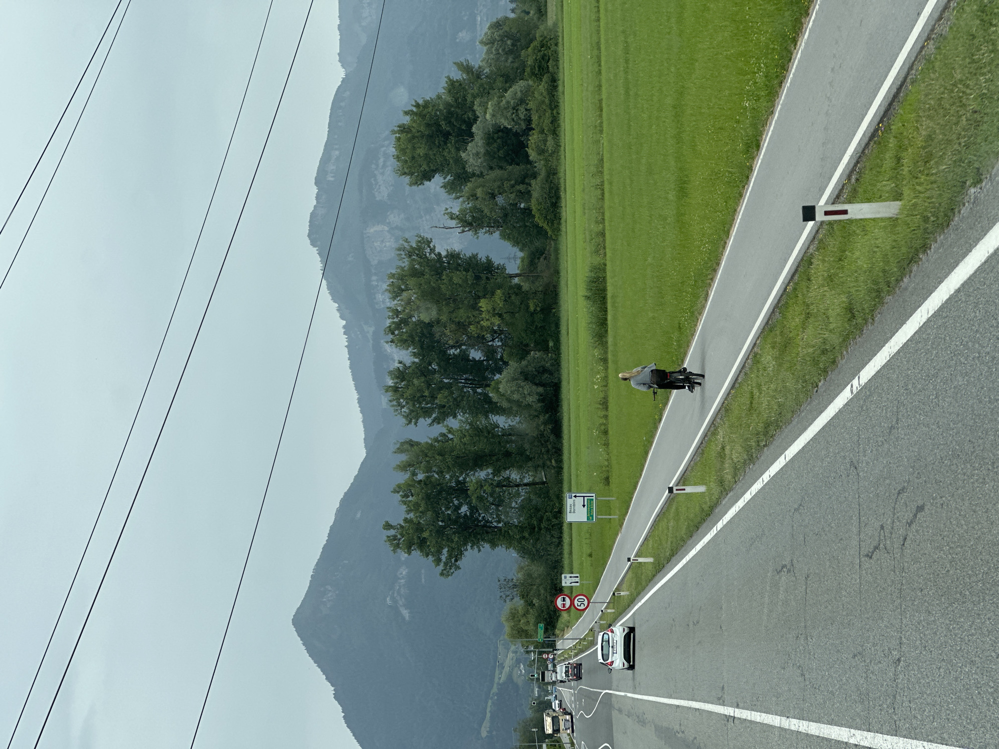 The image depicts a rural scene with a road, a bike path, a green field, and mountains in the background. A person is riding a bicycle on the bike path, moving away from the viewer. A white car and several trucks are visible on the road. Traffic signs indicating speed limits and directions are present along the road. The environment appears to be a cloudy day. The location is identified as Wolfurt, Austria.