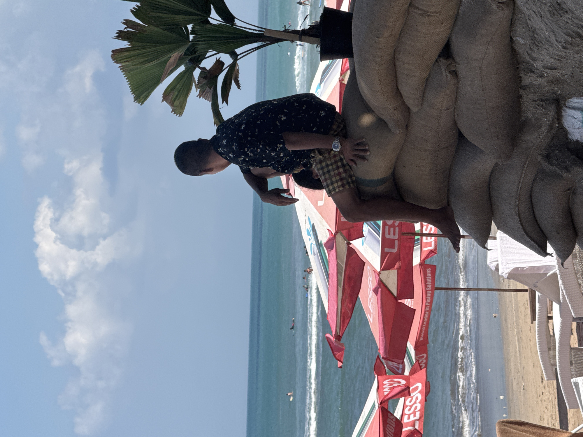 A man sits on a stack of burlap sacks on a beach in Kuta, Indonesia. He is wearing a short-sleeved patterned shirt and plaid shorts, and is looking out at the ocean. There are several red and white beach umbrellas in the foreground, and a few people can be seen surfing in the distance. The sky is blue with scattered clouds. The scene appears to be a sunny day, likely in the afternoon.