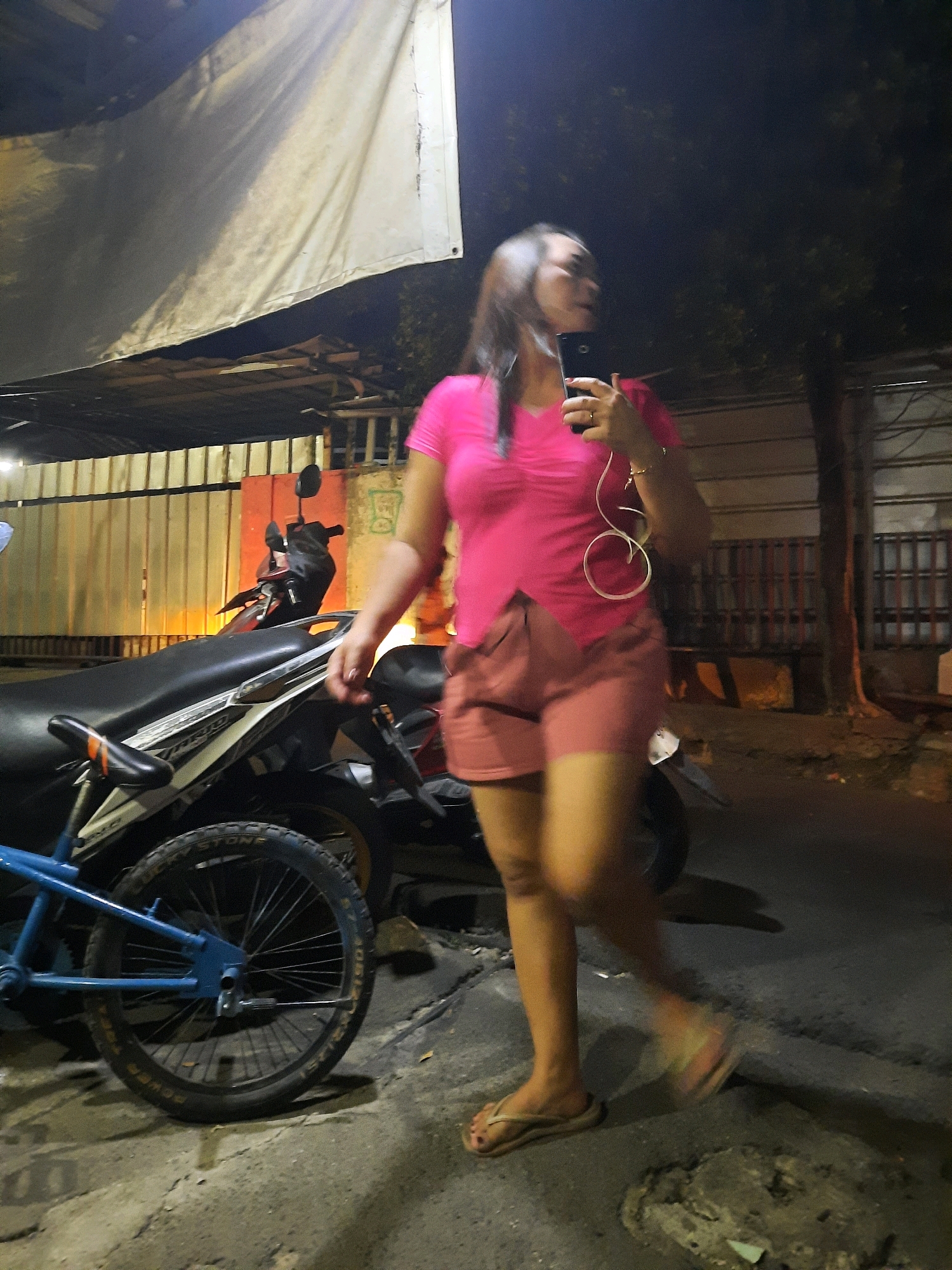 A blurred, low-light, full-body image shows a woman in a bright pink t-shirt and pink shorts walking on a cracked asphalt road at night. She is holding a black smartphone in her right hand and has white earbuds in her ears. The woman is wearing flip-flops on her feet. To her left, a blue bicycle is parked, and behind that, a motorcycle is partially visible. The background features a metal fence with orange and red accents, and a large white tarp is stretched overhead. The scene suggests a street in East Jakarta, Indonesia, likely a residential or commercial area given the presence of parked vehicles and buildings.