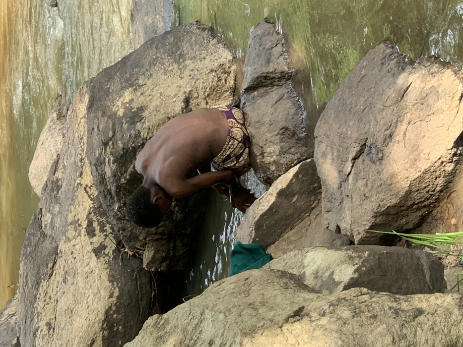 A young man is sitting on a large rock in a shallow stream. He is shirtless and wearing a pair of colorful patterned shorts. He is cupping his hands in the water, possibly washing them. He is sitting near the bank of the stream, surrounded by large rocks. The stream is flowing gently, and the water is a light green color. There is a green cloth lying on the ground nearby. The location is a small stream in the Egbeda area of Lagos, Nigeria, specifically at the coordinates 7.119° N, 5.362° E.