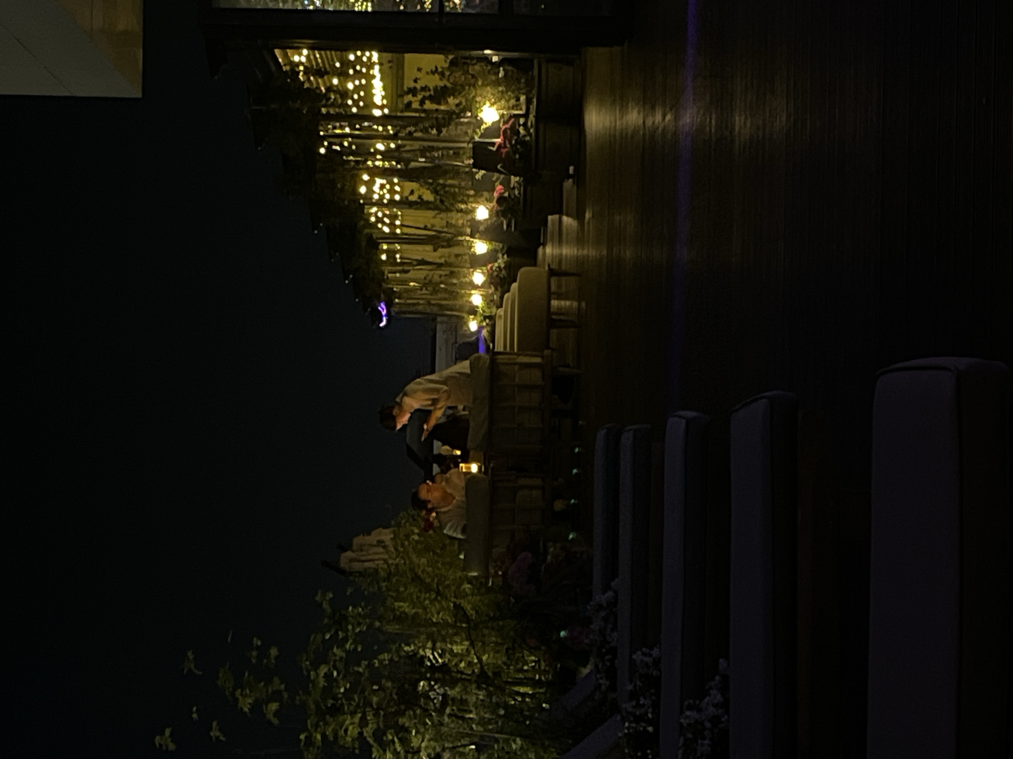 The image depicts an outdoor dining area at night, likely in Kazan, Russia. The scene is illuminated by string lights overhead, which are draped across the structure. Two people are seated at a table: one is wearing a light-colored top and the other is wearing a darker outfit. The dining area is situated on a wooden deck. There are green plants in the foreground and some pillars.