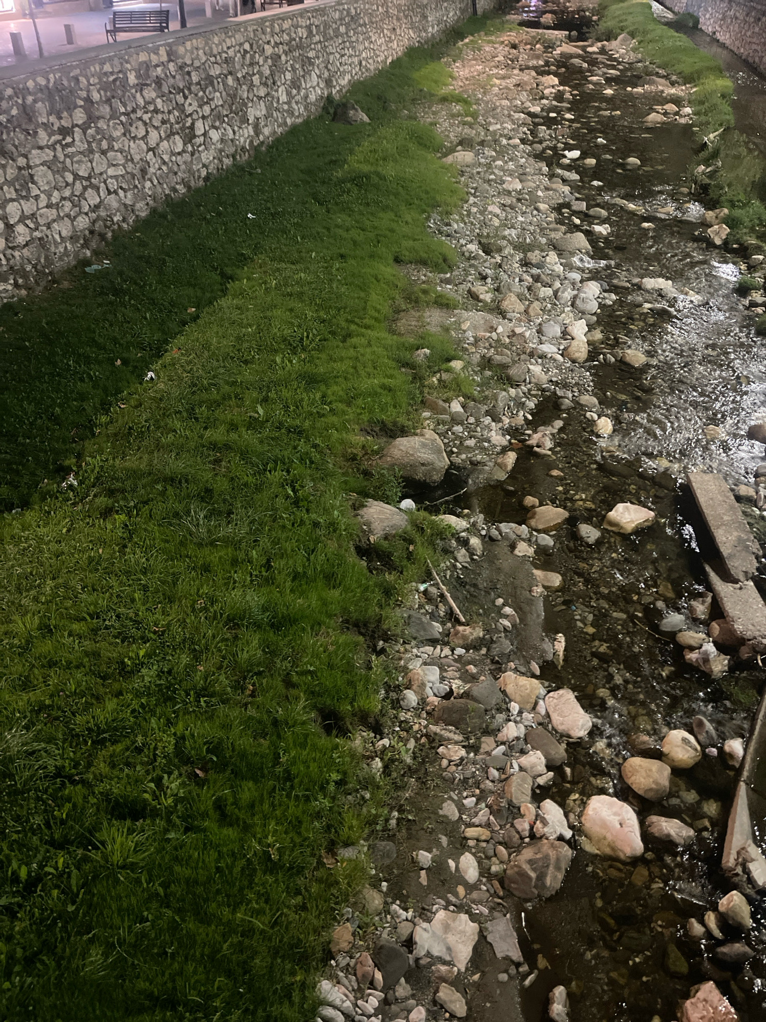 The image depicts a stone-walled embankment along a shallow, rocky stream in Prizren, Kosovo. The embankment, constructed from irregular, light-colored stones, runs along the left side of the frame and is topped with a paved walkway where two dark benches are visible. A strip of green grass borders the embankment and slopes down towards the stream. The stream itself is characterized by a bed of varied-sized, rounded rocks and pebbles, with clear water flowing over them. Some areas of the stream bed are exposed, while others are submerged in shallow water. The scene is illuminated by artificial lighting, casting reflections on the water and creating shadows. The overall environment suggests an urban or semi-urban setting with a natural water feature integrated into the landscape.