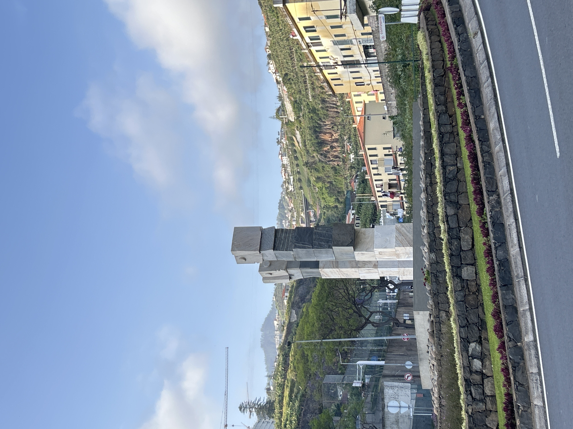 The image depicts a monument in Câmara de Lobos, Portugal, constructed of stacked stone blocks. A road runs parallel to the monument. The background includes a hillside with buildings. The sky is visible with scattered clouds. A construction crane is also present in the scene.