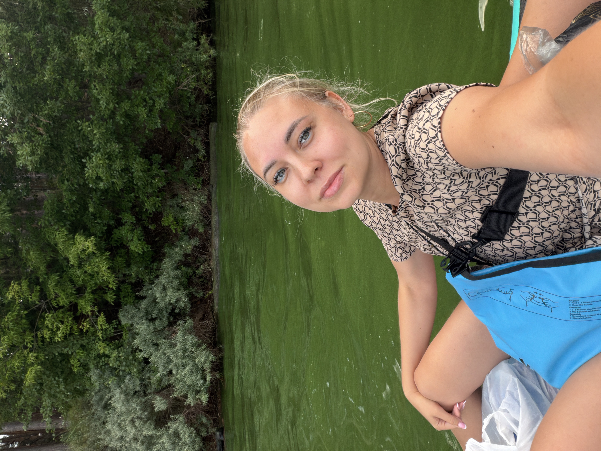 The image features a young woman with blonde hair and blue eyes, positioned on a body of green water, possibly a lake or river. She is wearing a patterned short-sleeved shirt and a blue dry bag with instructions printed on it. Her posture suggests she is sitting or kneeling, possibly on a boat or paddleboard.
To her left, dense green foliage forms a backdrop of trees and bushes, indicating a natural outdoor setting. The water's surface is textured with ripples and some white foam, hinting at movement. The lighting appears to be natural daylight, suggesting the photo was taken during the day. The woman's expression is relaxed and she is looking towards the camera.
The visible text on the blue dry bag provides instructions in multiple languages, including English, German, French, and Spanish, for closing the bag. The instructions are accompanied by simple line drawings. The context provided places this scene in Chaikovsky, Russia.