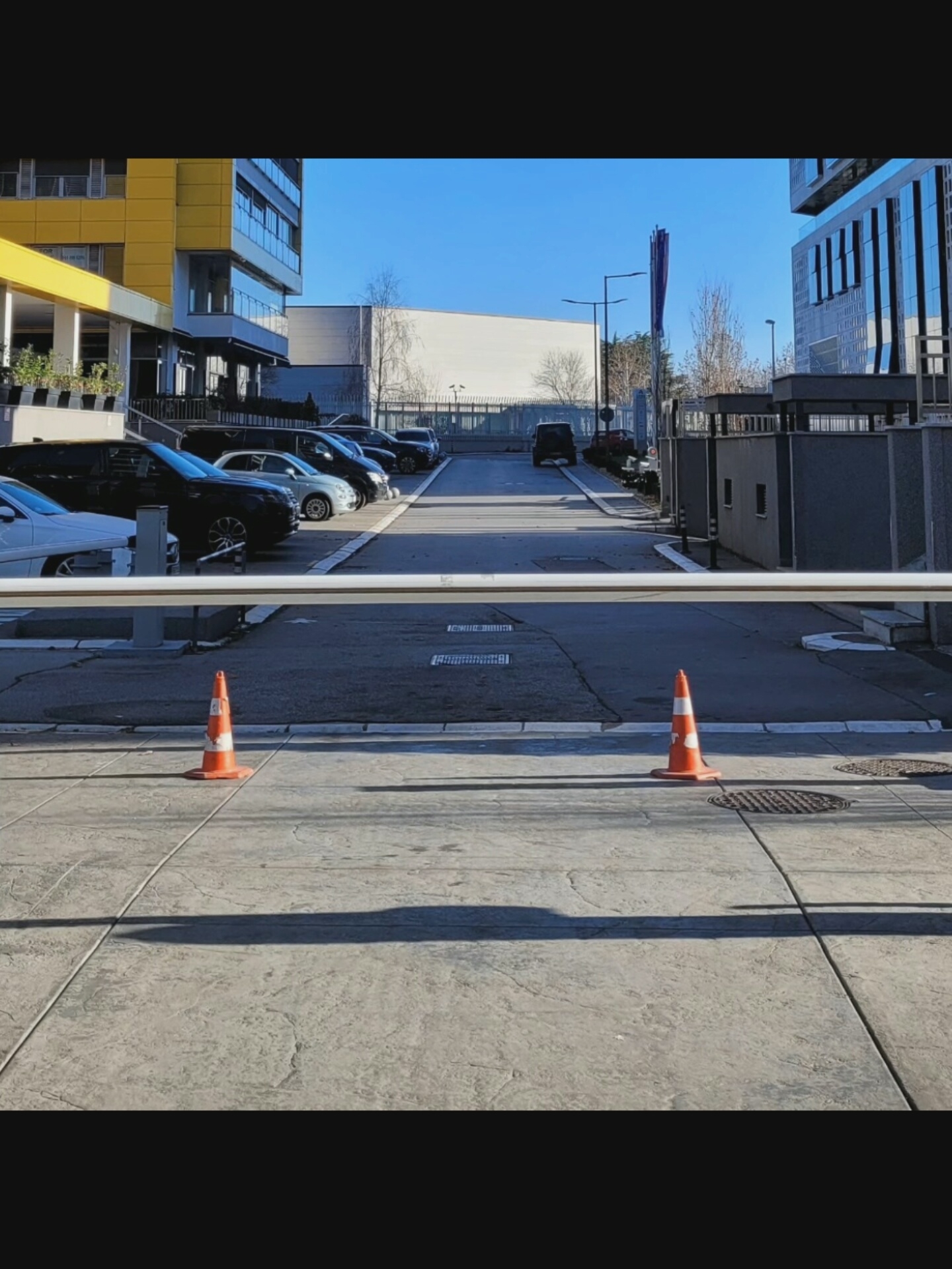 In Bielawska Wieś, Poland, a modern parking area is depicted under a clear blue sky. Two orange traffic cones are positioned on a grey paved surface, one to the left and one to the right, casting distinct shadows. A white horizontal barrier stretches across the mid-ground. Beyond the barrier, a row of parked cars is visible, including black SUVs and a silver hatchback. In the background, contemporary buildings with yellow and blue facades stand, along with a white rectangular structure and trees. A black Jeep is also present in the parking area. The scene appears to be a secure, organized, and well-maintained urban space.