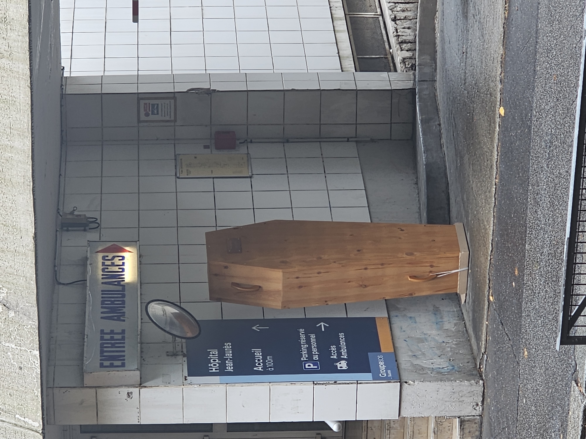 The image depicts an exterior entrance area for a building, likely a hospital, in Paris, France. A light-colored wooden coffin is prominently displayed, standing vertically on a paved ground surface, positioned against a tiled wall. The coffin has a simple design with wooden handles.
The tiled wall, which forms the left side of the scene (after a 90-degree clockwise rotation for proper viewing), features several informational signs. A rectangular illuminated sign reads "ENTREE AMBULANCES" (Ambulance Entrance) with an upward-pointing arrow. Below it is a large blue directional sign with multiple entries: "Hôpital Jean-Jaurès" (Jean-Jaurès Hospital), "Accueil à 100m" (Reception at 100m) with an arrow, "Parking réservé au personnel" (Staff parking) with a 'P' icon and an arrow, and "Accès Ambulances" (Ambulance Access) with an ambulance icon and an arrow. The logo "Groupesos Santé" is also visible on this sign. A convex security mirror is mounted on the wall near the ambulance entrance sign. Other minor details on the tiled wall include a red box (possibly a fire alarm or electrical panel) and some exposed wiring.
The ground is a dark, paved surface, resembling asphalt, with a slightly different texture on the right side, where a metal railing is partially visible. The overall setting appears utilitarian, with the tiles and concrete structures showing some wear.
No people are visible in the image. The scene is illuminated by natural daylight, indicating it was captured during the day. The presence of the coffin in this context is unusual and striking.