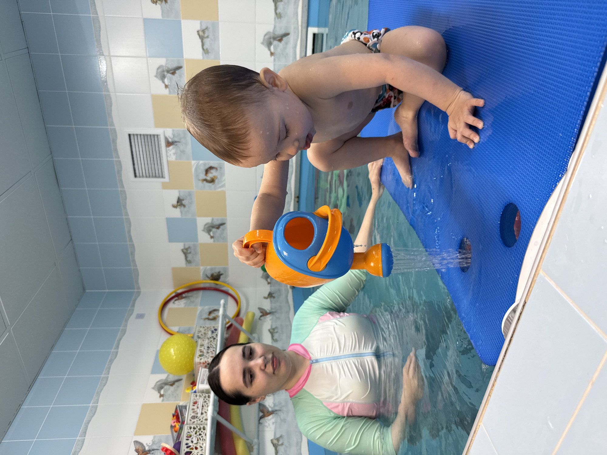 A small child in swim trunks sits on a blue rectangular floating mat in a swimming pool, pouring water from an orange and blue watering can onto the mat. A woman in a white, pink, and light green swimsuit stands in the water, holding her hands towards the child. The pool is surrounded by light blue and white tiled walls. A vent is on the upper left wall. The walls are decorated with tiles patterned with birds. A metal rack with yellow and red hoops and a yellow ball sits in the background. The location is Rusanovo, Russia.