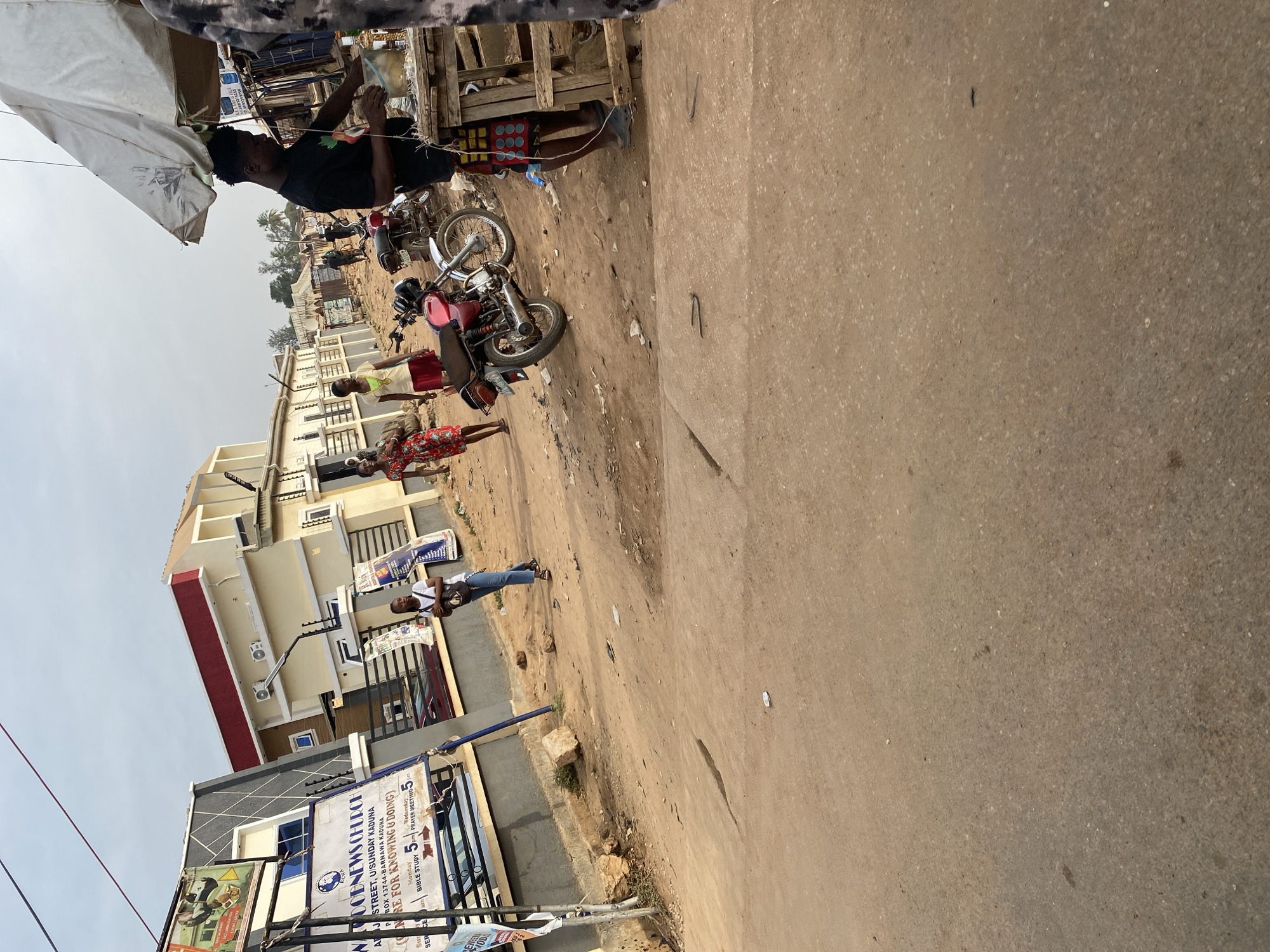 This is a candid, eye-level photograph capturing a street scene, likely in Sabon Tasha, Kaduna, Nigeria, during the daytime under a clear sky. The image portrays daily life in a bustling local community.
In the foreground on the left, a man in a dark t-shirt is seen under a white canopy or umbrella, attending to what appears to be a street stall or vendor setup. He is in the process of pouring liquid into a clear container. Adjacent to the stall, two motorcycles are parked on the unpaved ground: one is a prominent red color, and the other is lighter-colored. Wooden crates and other miscellaneous items are scattered around the stall area.
Further into the scene, several individuals are visible. A woman in a red and white patterned dress walks in the mid-ground, and another person in a yellow top stands nearby. Towards the left of the frame, a person wearing a white t-shirt and blue jeans is standing near a building. The ground is a mix of dirt, gravel, and unpaved sections, leading to what appears to be a wider, paved road on the right.
The background features several multi-story buildings, characterized by light-colored facades with occasional red or dark accents. A distinctive sign for "DOBNEWS CHURCH" is prominently displayed, indicating its location as "U/SUNDAY KADUNA" and "BOX 13744-BARNAWA KADUNA." The sign also advertises service times: "Monday 5pm" and "Wednesday BIBLE STUDY PM PRAYER MEETING 5." Power lines crisscross the upper portion of the frame, and distant trees are visible under the bright sky, completing the impression of a typical urban or semi-urban Nigerian setting.