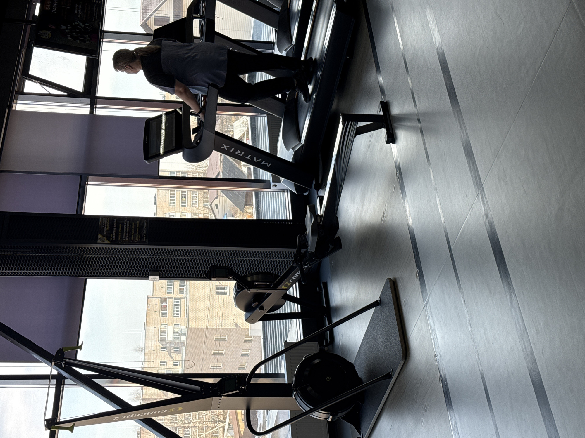 The image captures a gym interior in Votkinsk, Russia. A woman is seen on a Matrix treadmill, exercising. A Concept 2 rowing machine is also visible in the foreground. The gym has large windows offering a view of buildings outside.
