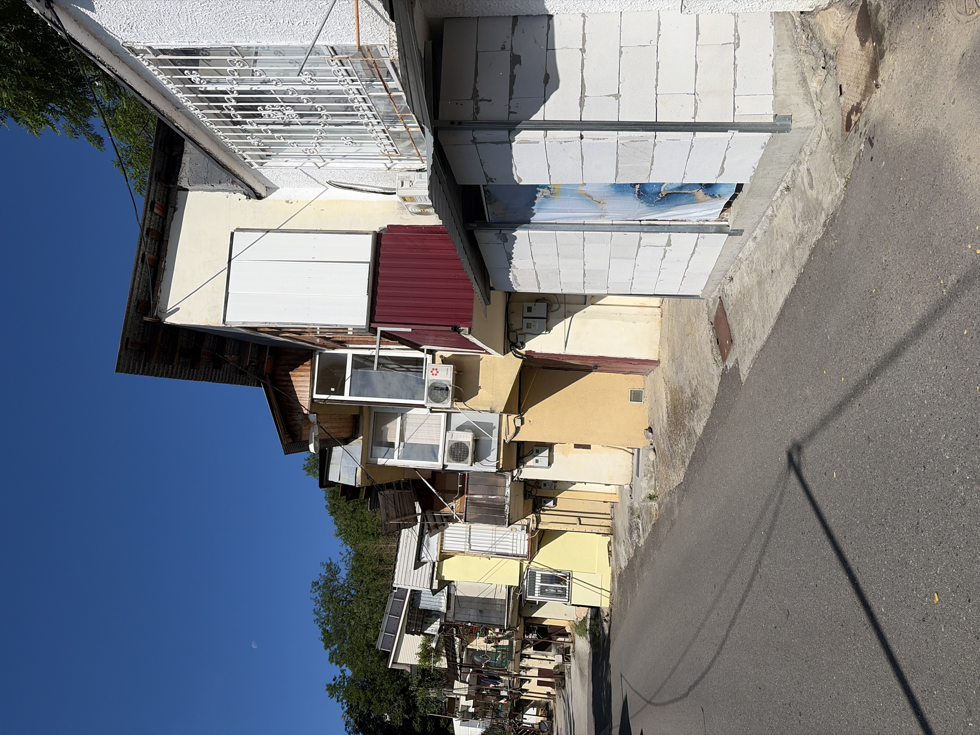 This image captures a street scene in the city of Sochi, Russia, on a clear, sunny day. The scene depicts a narrow asphalt road bordered on the right by a continuous line of multi-story residential buildings, and on the left by a steep, tree-covered hillside.
The buildings exhibit a varied and somewhat informal architectural style, characterized by numerous additions and modifications. Facades feature a mix of painted surfaces (yellow, beige, white), exposed concrete blocks, and corrugated metal. Several structures incorporate enclosed or open balconies, often equipped with external air conditioning units. Some upper levels display solar panels mounted on rooftops or balcony extensions. Windows vary in style; one prominent window on a white building in the mid-right features elaborate white security bars. The building closest in the foreground on the right has a partially completed or renovated section constructed from white aerated concrete blocks, covered by a corrugated metal awning, with a blue and white patterned tarp or poster obscuring an opening.
The asphalt road shows visible tire marks and is partially illuminated by long shadows cast by unseen overhead lines or poles. The ground adjacent to the buildings consists of concrete pathways and bare earth, where two distinct metal manhole covers are visible.
The background is dominated by a densely forested hill, indicating the mountainous or hilly terrain characteristic of Sochi. The sky above is a clear, deep blue, with a faint crescent moon discernible in the upper left quadrant. No individuals are visible in the scene. The overall lighting suggests bright midday sun, casting strong shadows.