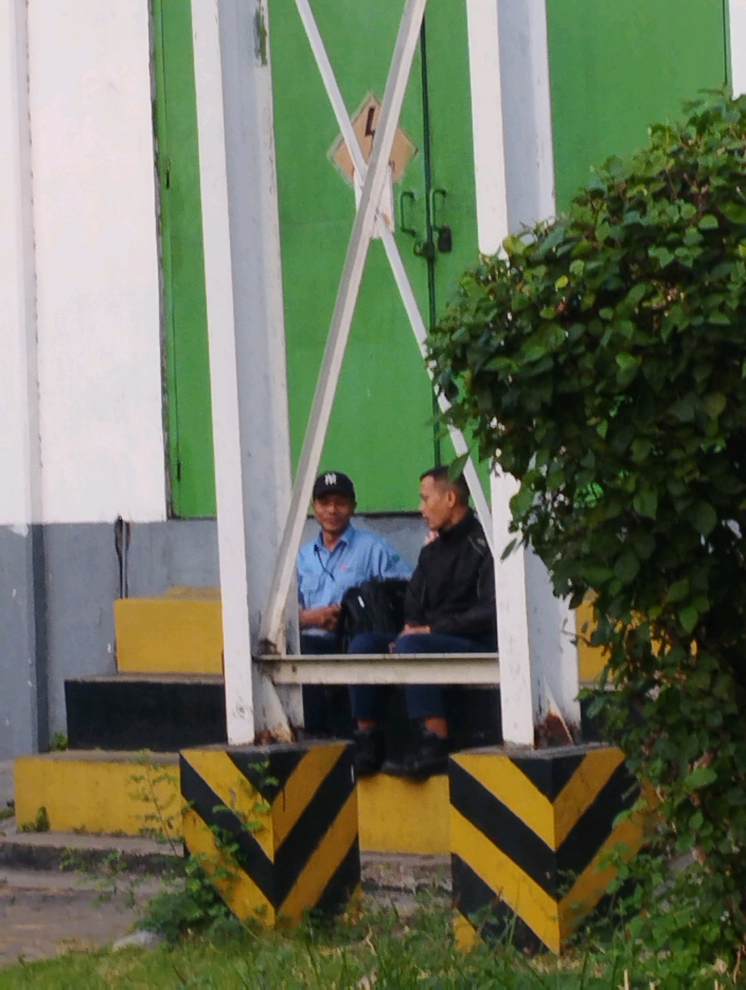 Two men are seated on yellow steps outside a green building. The man on the left wears a blue shirt and a black baseball cap with a white logo, and he is smiling. The man on the right wears a black jacket and dark pants, and he is looking to the left. A black backpack is visible on the ground next to him. The building has a white frame structure in front of it, and green doors with a yellow diamond shape sign with a black symbol are visible on the doors. Yellow and black striped bollards are present on the ground in front of the steps. Lush green foliage partially obscures the right side of the image. The scene appears to be in an outdoor, possibly industrial or commercial, area in Kramat Temenggung, Indonesia.