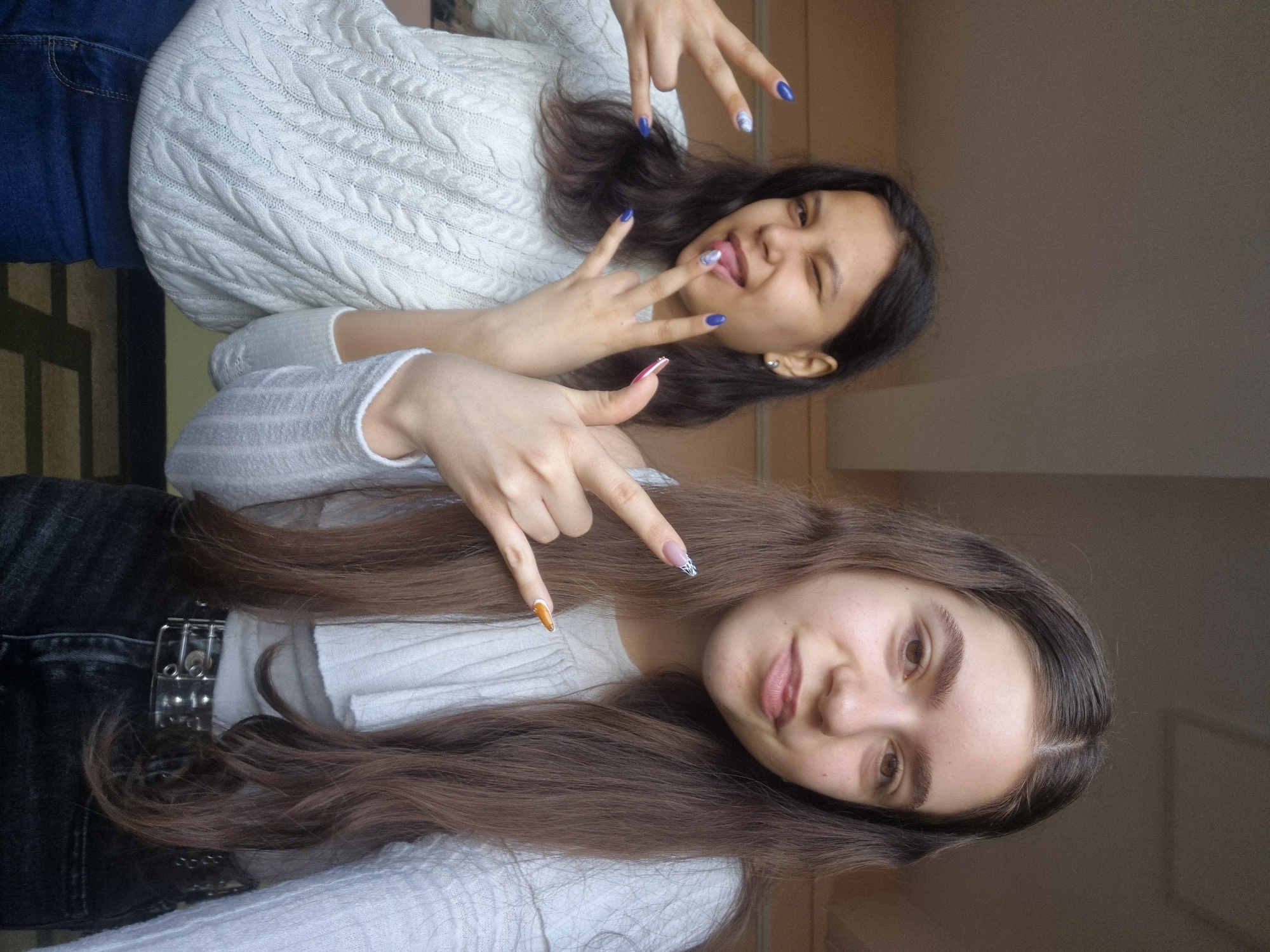 Two young women are posing for a photograph indoors. The woman in the foreground has long brown hair and is wearing a white sweater. She is making a hand gesture with her fingers. The woman in the background has dark hair and is wearing a white sweater and blue jeans. She is sticking out her tongue and making a hand gesture. The location is not discernible.