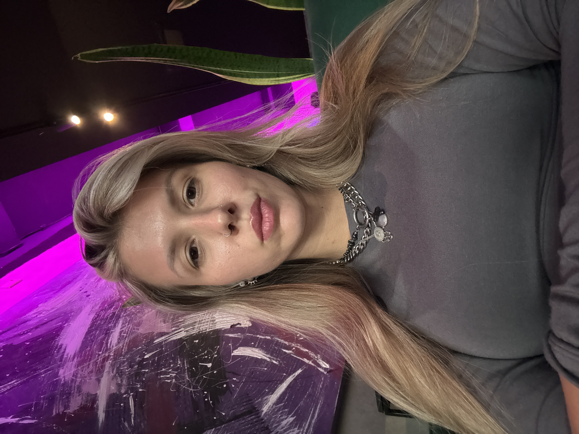 A young woman with blonde hair and a nose ring poses for a selfie indoors. She is wearing a gray long-sleeved shirt and a chunky silver necklace with charms. The background features a large abstract painting in shades of purple and white, and a tall snake plant. The lighting is dim with bright purple and warm yellow lights illuminating the scene.