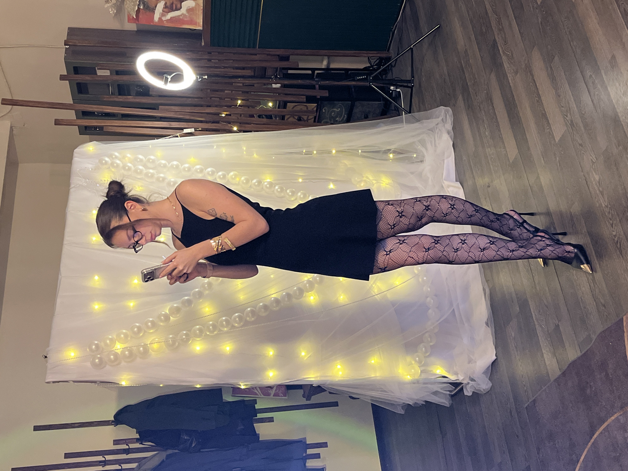 A woman stands in front of a backdrop adorned with strings of pearls and fairy lights. She is wearing a black dress, patterned stockings, and black high heels with gold accents. She holds a smartphone, looking down at the screen. The location appears to be indoors, with a wooden floor and a wooden structure behind the backdrop. The city of Saint Petersburg, Russia, is not evident from the image.