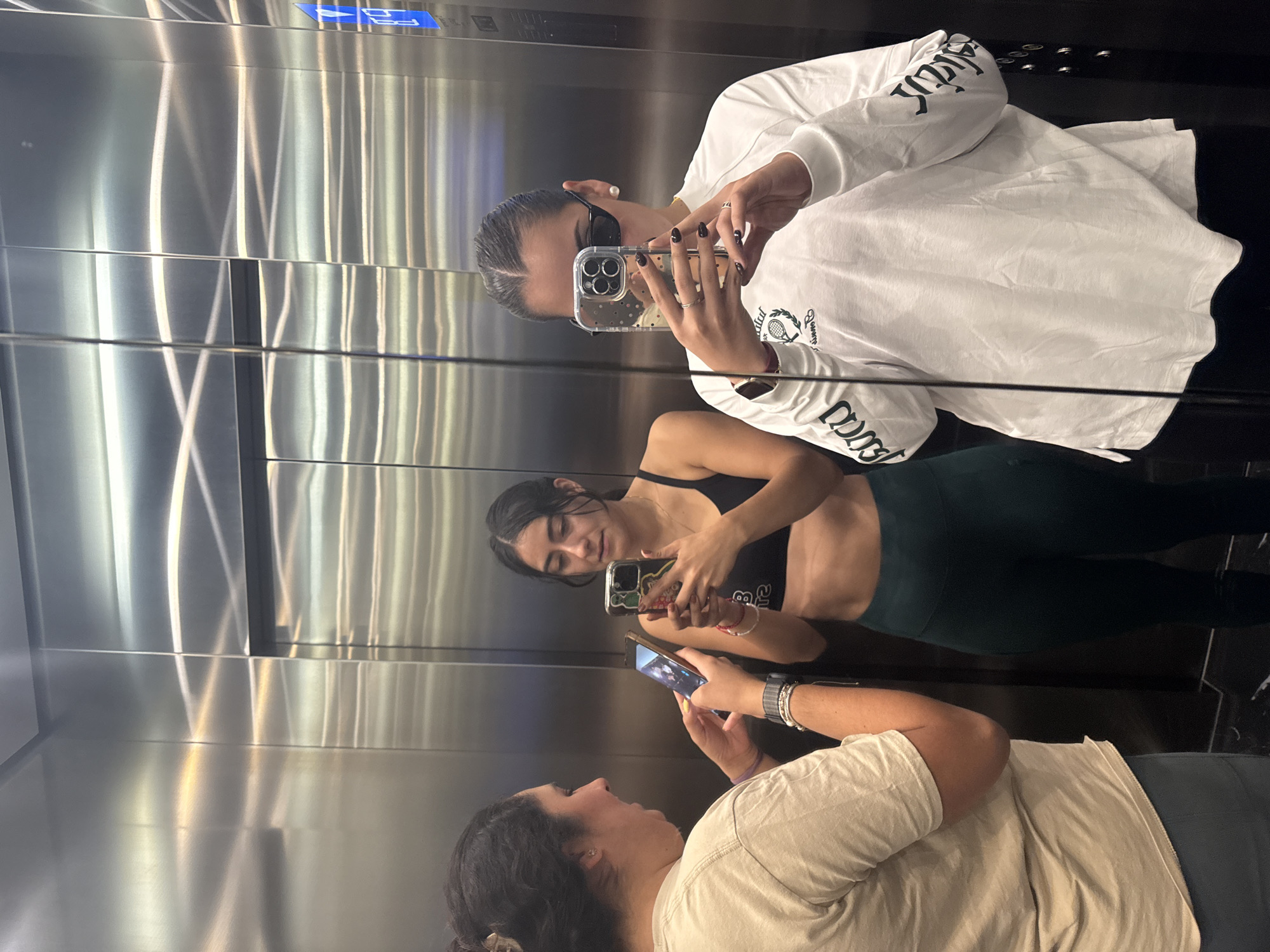 Three women are in an elevator with stainless steel walls and a black floor. The woman at the top of the image wears a white long-sleeved shirt with black letters on the sleeves and takes a photo with her smartphone. The woman in the middle wears a black sports bra and dark green leggings, also using a smartphone to take a photo. The woman at the bottom of the image wears a light-colored t-shirt and gray bottoms, looking at her smartphone. The elevator is in Puebla, Mexico.