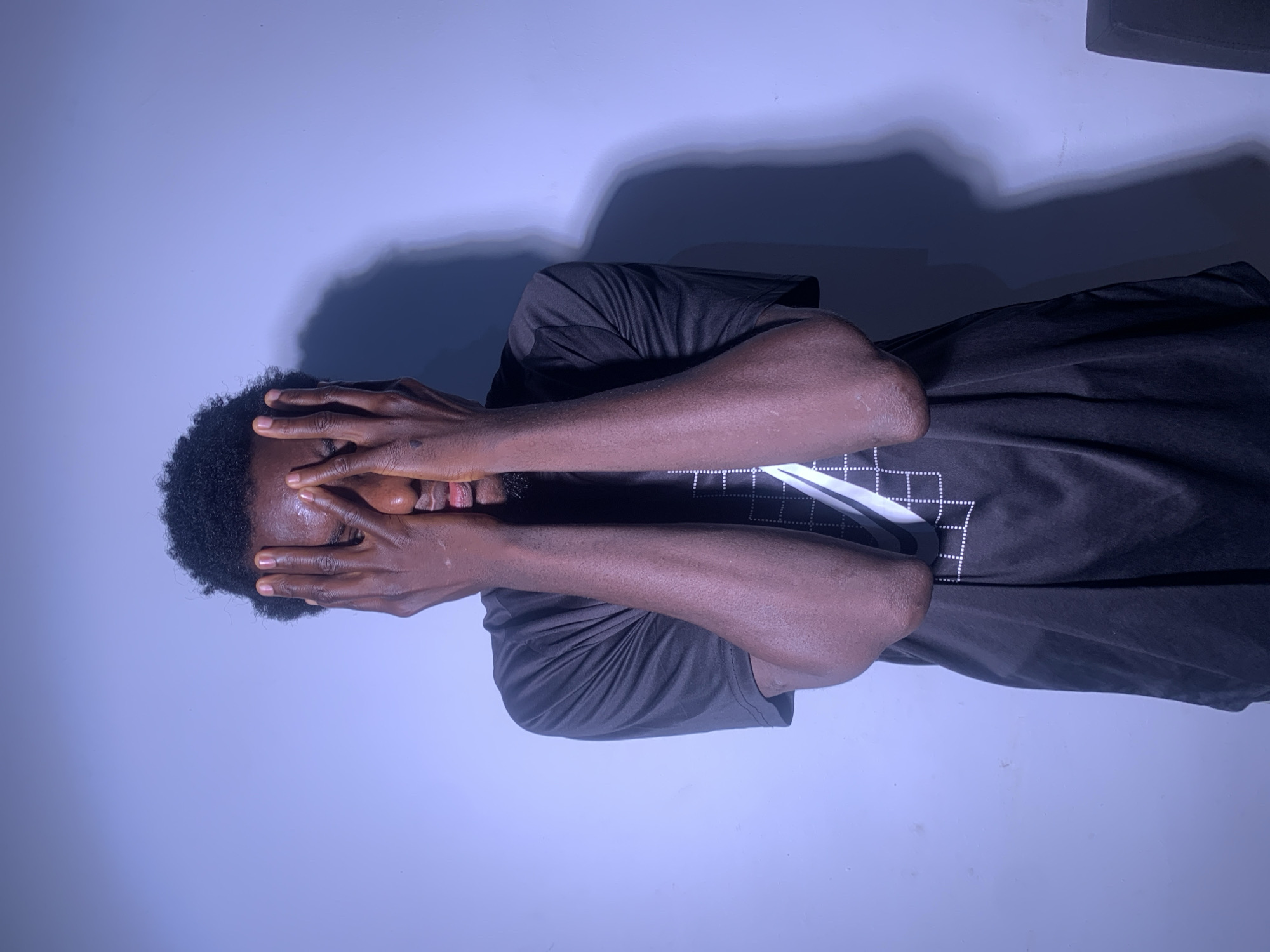 A young Black male stands against a light purple wall, partially obscuring his face with his hands. He is wearing a black t-shirt with a white graphic design on the front. His arms are bent at the elbows, and his fingers are interlaced in front of his eyes. A shadow of his head and shoulders is cast on the wall to his right. The lighting appears to be artificial, casting a blueish tint on the scene. The background is a plain, smooth wall with a slight texture visible upon close inspection.