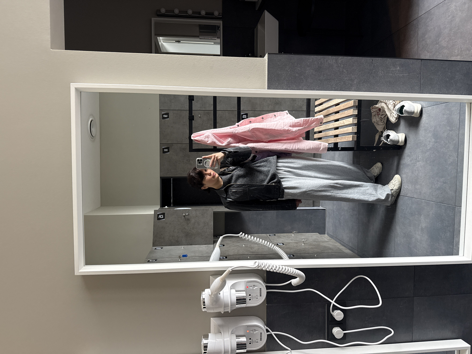 The image shows a reflection in a mirror within a bathroom or changing room, potentially located in Berlin, Germany. The mirror frame is white and reflects a person holding a smartphone, taking a selfie. The person is wearing a grey sweater, grey sweatpants, and a black leather jacket. A pink shirt hangs on a wooden rack behind the person. On the rack sits a pair of sneakers and a second pair on the floor near the person. The background of the reflection shows grey lockers with numbering on them, indicating individual compartments. On the wall outside the mirror's reflection, two white hair dryers are mounted, with their cords and electrical outlets visible. The wall is light beige and the floor is made of dark grey tiles. Above the mirror, a partial view of another mirror and cabinetry is visible.