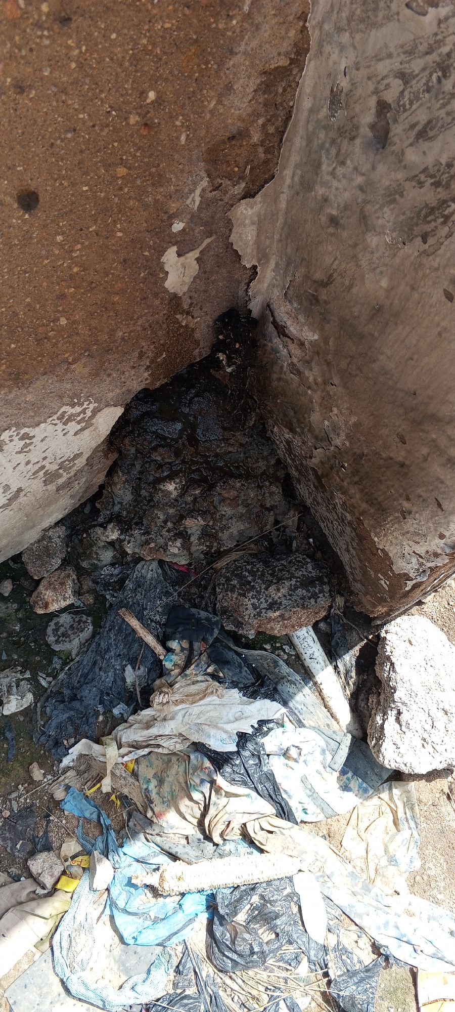 The image shows a pile of refuse in a corner formed by two concrete structures in Jalingo, Nigeria. The refuse includes plastic bags, cloth scraps, rocks, and what appears to be stagnant water or mud. The concrete structures show signs of wear and tear, with some areas of exposed aggregate and peeling paint or cement. The ground is a mix of dirt and debris. The overall scene depicts an unmaintained and unsanitary environment.