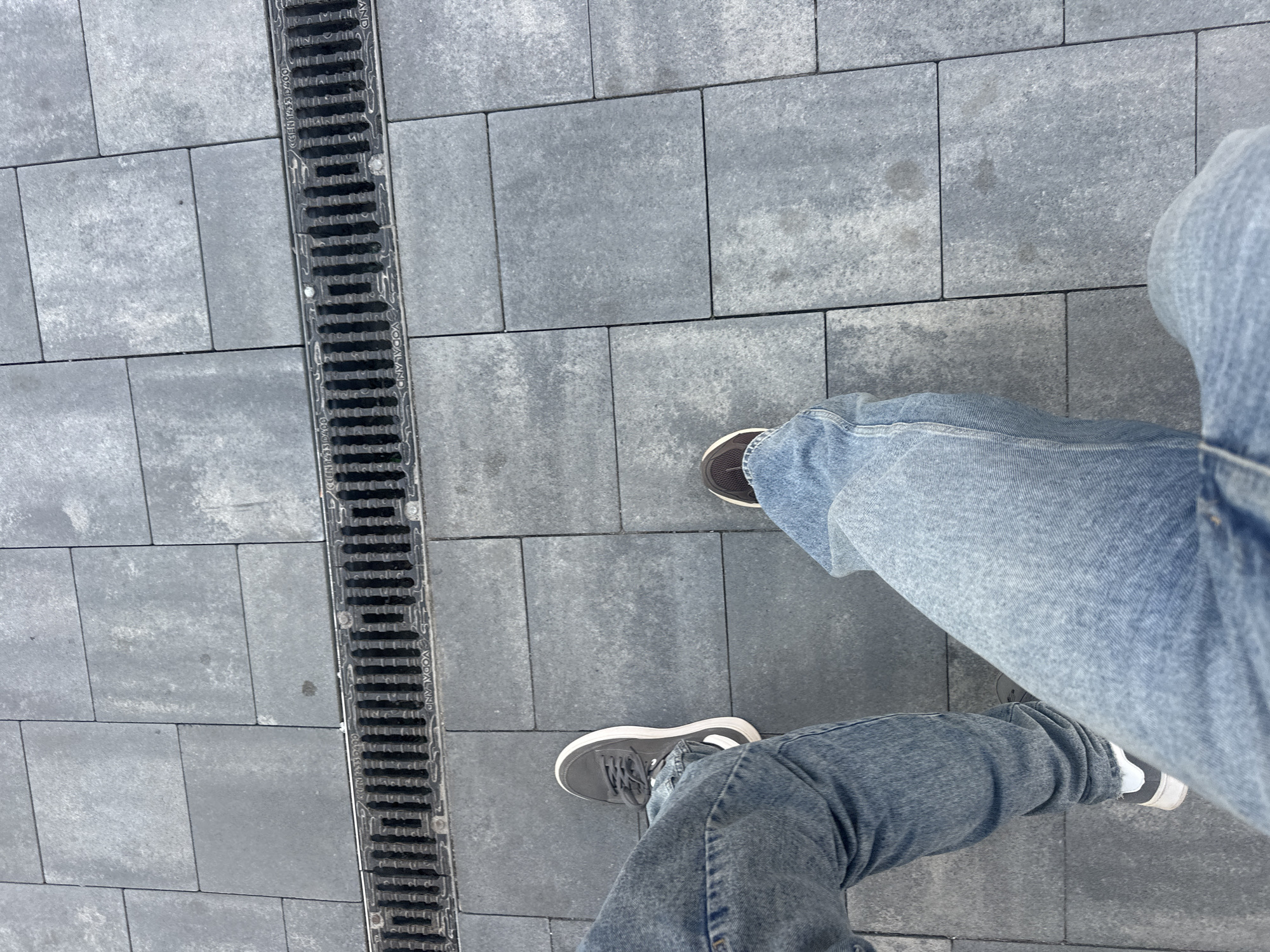 The image depicts a section of pedestrian pavement, likely in an urban setting. The pavement is constructed from grey rectangular paving stones. A linear drainage channel runs vertically on the left side, featuring parallel metal grates. A person's legs, clad in light blue denim jeans, are positioned on the pavement. The individual is wearing sneakers; the left foot is shod in a grey sneaker with a white stripe, while the right foot appears to be in a brown sneaker. The scene appears to be in the city of Ternopil, Ukraine. The photograph is taken from a high-angle perspective.