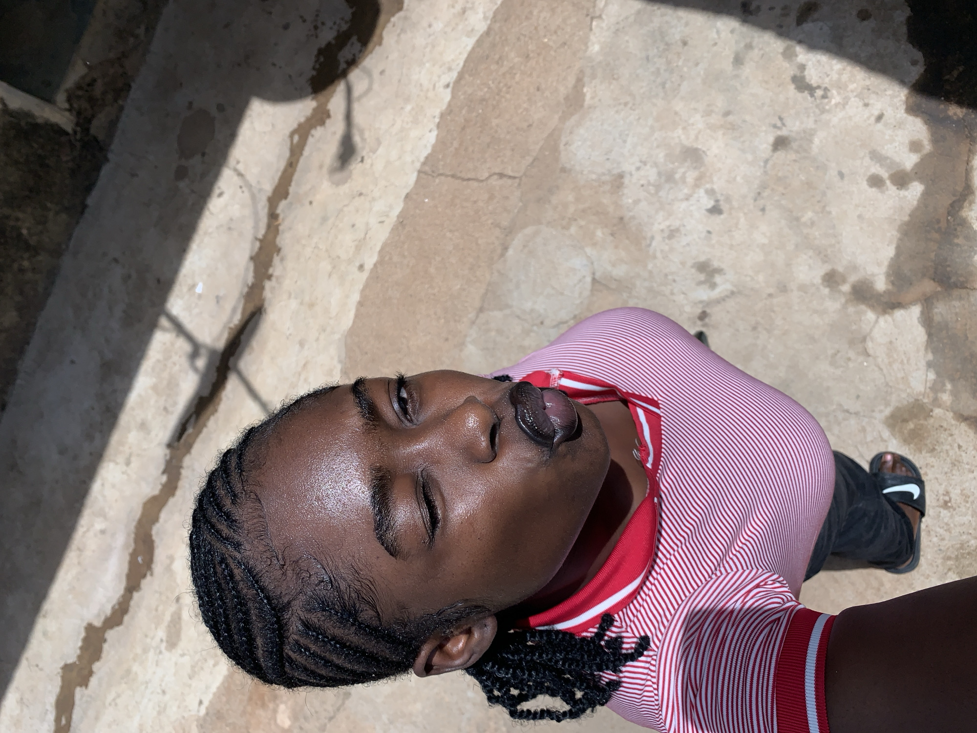 The image captures a young person, likely female, from an overhead perspective, looking upwards towards the camera. The subject has dark skin, braided hair styled in cornrows, and appears to be in their late teens or early twenties. They are wearing a red and white striped short-sleeved top, dark pants, and a black slide sandal with a white Nike logo on the strap is visible on their right foot.
The person's expression is playful, with their tongue slightly out and their left eye squinted or winking, while the right eye is more open. Their face shows a noticeable sheen, possibly from sweat or oil, indicative of warm weather.
The setting appears to be outdoors, with the person standing on rough concrete or paved ground. The ground shows cracks and some dark stains. Strong shadows are cast across the ground, suggesting bright sunlight, indicating it is daytime, likely midday or early afternoon. The overall atmosphere is casual and relaxed. The location context is Rafin Par, Nigeria.