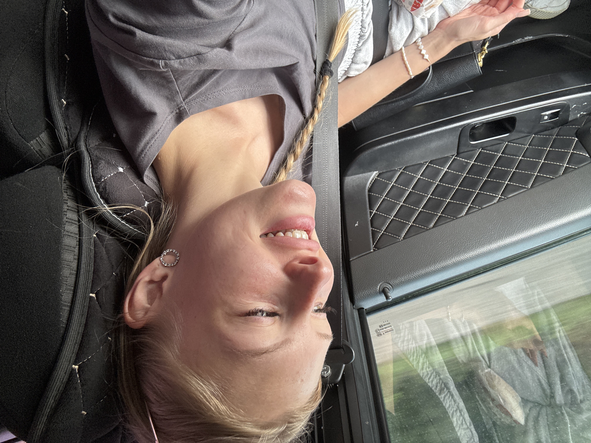 The image shows a young blonde woman with her hair in a braid, wearing a grey t-shirt and a pearl bracelet. She is sitting in a car, with the car's interior visible, including the door panel with diamond stitching and a seatbelt. Through the window, a blurry green landscape is visible, suggesting the car is in motion. The woman is looking out the window and smiling. The location of Шумиха, Russia is not discernible from the image itself.