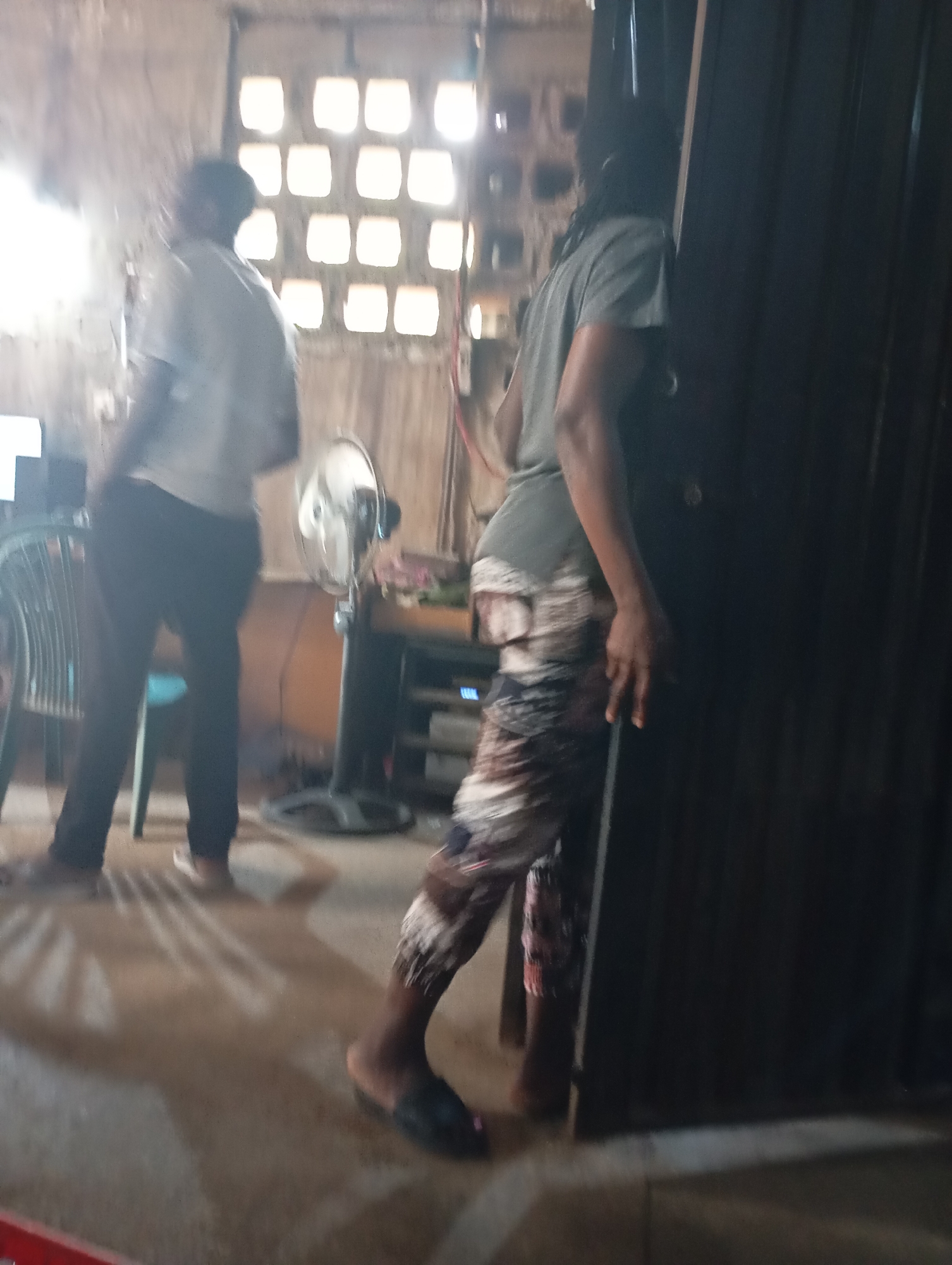 In the city of Shendam, Nigeria, a dimly lit room is depicted with two individuals. The individual on the left, wearing a light-colored t-shirt and dark pants, is standing with their back to the viewer. To the right, another person, in a gray t-shirt and patterned pants, is facing away from the camera, partially obscured by a dark, tall object, possibly a door or partition. In the background, a fan stands next to a shelf with electronics, and a window with multiple square panes allows light to enter the room. Shadows are cast on the floor, suggesting the light source.