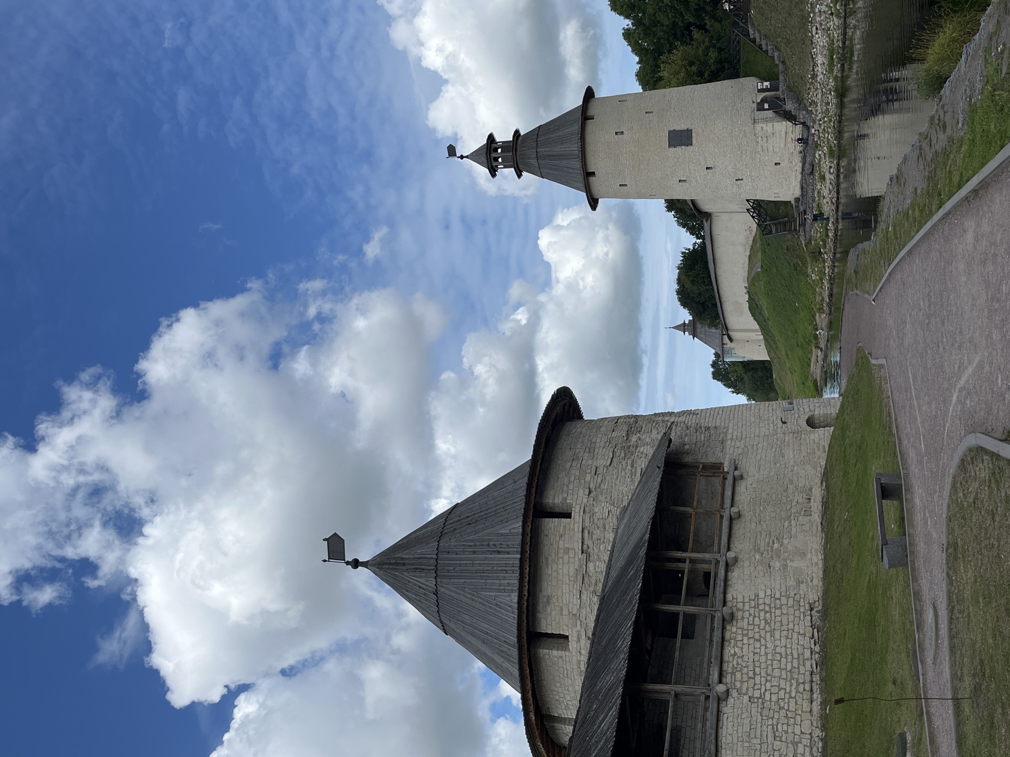 This is a vertical, eye-level, medium shot of the Pskov Kremlin in Pskov, Russia, under a bright blue sky dotted with white, puffy clouds. In the foreground, a paved path curves through a green lawn. To the left is a large, conical-roofed stone tower of the Kremlin with wooden upper walls. A wooden walkway with railings runs along the side of the tower. To the right, a lighter, slender stone tower with a similar conical roof stands taller. Stairs lead up to its entrance. A body of dark water, reflecting the sky and towers, runs along the right side of the path. In the background, the long white wall of the Kremlin stretches into the distance, flanked by lush green trees. The overall impression is one of historical grandeur and natural beauty.