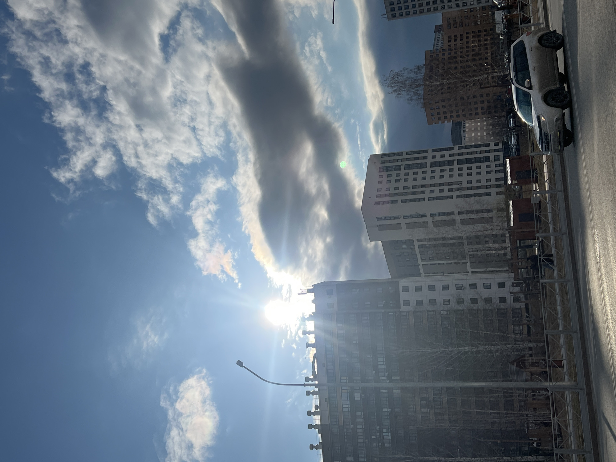 This image captures a bright, sunny daytime scene in an urban environment, likely a city in Tyumen, Russia, as indicated by the context. The sun is prominently featured, shining brightly from the left side of the frame, creating a strong sunburst effect and lens flare as it peeks through scattered clouds. The sky is a clear blue, interspersed with both fluffy white clouds and longer, darker cloud formations that partially obscure the sun.
Several modern high-rise buildings dominate the right and central portions of the image. These buildings vary in color, with some appearing light grey or white and others a darker brown, all featuring numerous windows. In the foreground on the lower right, a paved street or road is visible, alongside a metal fence. A white SUV is parked or moving on this street. A black, curved streetlight pole stands in the lower-left foreground. Bare deciduous trees are visible between the buildings, suggesting late winter or early spring.
There are no people visibly present, and no specific activity or event is taking place, presenting a tranquil urban landscape. The overall impression is one of a clear, bright day in a modern city. No discernible text is visible anywhere in the image.