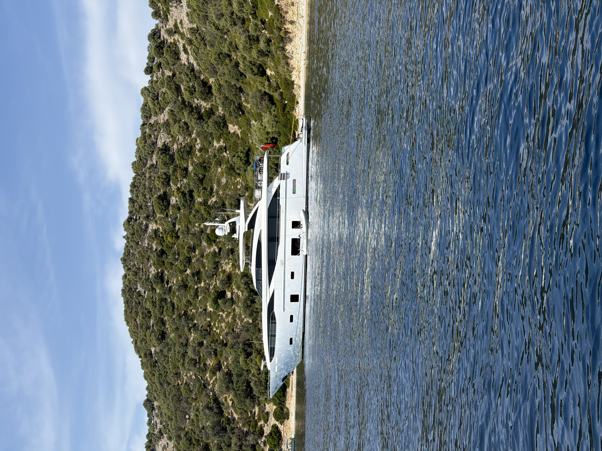 A large, white motor yacht is anchored in a calm, blue body of water, close to a shore lined with a rocky beach and a densely wooded, green hillside. The yacht is the primary subject, taking up the majority of the mid-ground and extending from the left to the right side of the frame. Its sleek, modern design features multiple decks and large windows. A small, inflatable dinghy is attached to the stern of the yacht. On the shore, a few small, dark birds are visible near the waterline. The hillside behind the yacht is covered in lush green vegetation and scattered rocks, with the blue sky and sparse white clouds above. The overall scene suggests a peaceful, scenic location. The Municipal Unit of Kastos is an island municipality in Greece, and this image likely depicts a bay or cove on the island, given the natural landscape and the presence of a yacht.