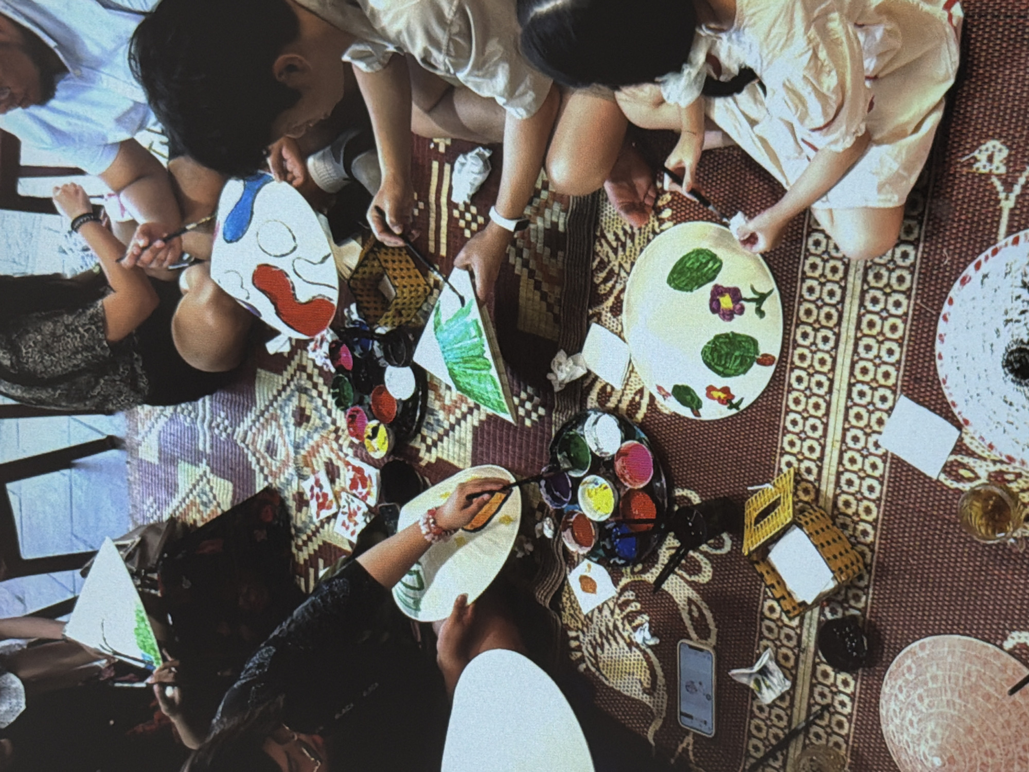People are seated on a rug, painting objects that appear to be non la (Vietnamese conical hats). Painting supplies, including palettes of paint, are scattered around the rug. The location is likely in Đà Nẵng, Vietnam.