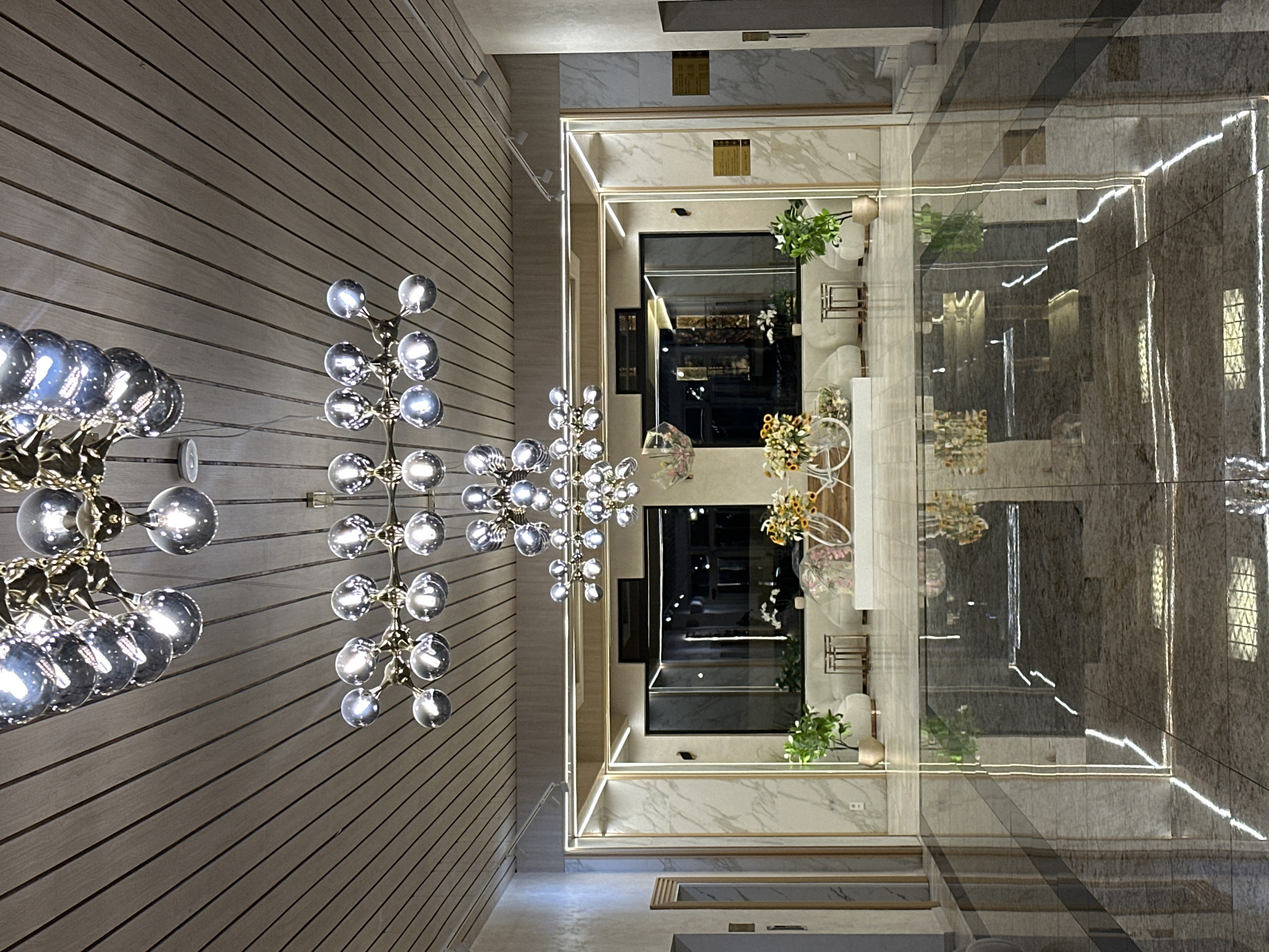 The image shows an interior space featuring modern lighting fixtures and architectural elements. Dominating the left side of the image is a wall with a light wooden texture. Three globe-shaped chandeliers, with their bulbs radiating light, adorn the wall.
In the center, a squared corridor or hall is framed with a marble-like material. This area features reflective flooring, enhancing the light and creating mirrored images of the scene. The walls within this area exhibit displays of greenery and decorative vases, positioned next to seating arrangements.
The space suggests a contemporary design, with a focus on light and reflection, indicative of a sophisticated interior. The location, Veliko Tarnovo, Bulgaria, is not visually discernible from the image alone.