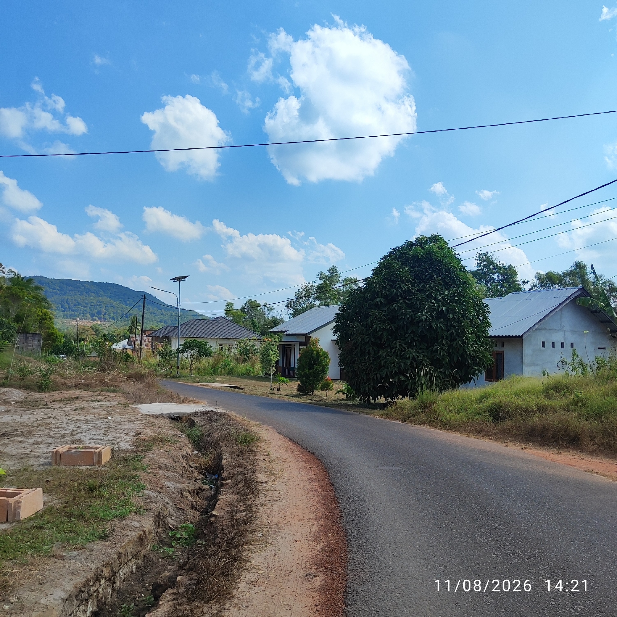 The image depicts a street scene in a rural area, likely the city of Pembaharuan, Indonesia, under a bright blue sky with scattered white clouds. The road, paved with asphalt, curves through the landscape. To the left of the road, the ground is rough and dry with some patches of grass and a small ditch. Two brick-like objects are visible on the left foreground. Further back, on the left, a wall made of what appears to be cinder blocks stands. Beyond this, a hill covered in dense green foliage dominates the background, with a few scattered houses visible at its base.

On the right side of the road, there are two houses with simple, gray metal roofs. The house closer to the foreground is white with a few small square windows on its side. A large, dense green tree stands to the right of this house. A second house is visible further down the road on the right, also with a gray metal roof. Various trees and vegetation are scattered throughout the scene. A solar-powered street light is positioned on a pole near the houses. The overall impression is a quiet, semi-developed rural environment. The date and time "11/08/2026 14:21" are superimposed on the lower right corner of the image. No people are present in the scene.