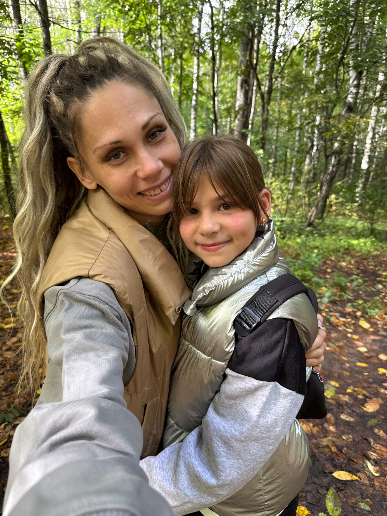 In Rusanovo, Russia, a woman and a young girl are captured in a selfie amidst a lush forest. The woman, with her blonde hair pulled back in a high ponytail, sports heavy eye makeup and a bright smile. She's wearing a light grey hoodie underneath a tan vest. Beside her, the girl, with brown hair and rosy cheeks, smiles warmly, her head resting on the woman's shoulder. She's dressed in a metallic green puffer jacket layered over a grey hoodie with black accents. The background is a vibrant forest scene, with tall trees and fallen leaves carpeting the ground. The overall atmosphere is one of happy togetherness in a natural setting.