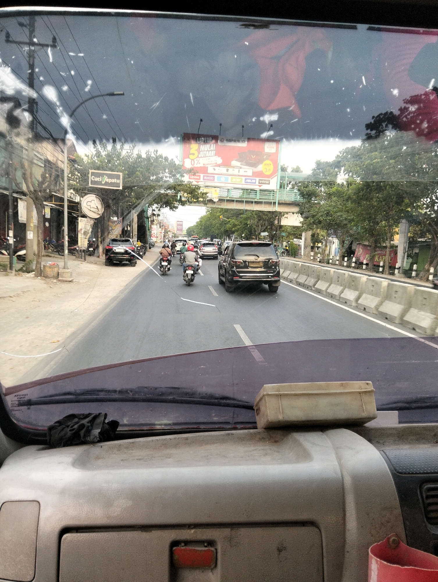 The image, taken from inside a vehicle, provides a view of a street scene in Semarang, Indonesia. The perspective is looking forward through the windshield, which is somewhat dirty, with several white streaks and specks visible.
In the foreground, the dashboard of the vehicle is visible, appearing dusty. A light-colored plastic container is placed on top of a section of the dashboard. To the left, a black cloth or rag is also resting on the dashboard.
The road ahead is a multi-lane asphalt road with a white line marking the lanes. Traffic is present, consisting primarily of motorcycles and a few cars. Several motorcycles are being ridden by people wearing helmets. One black SUV is prominently featured in the right lane, moving away from the camera. On the left side of the road, there are buildings, trees, a lamppost, and a sign that reads "Cuci Sepatu" (Shoe Wash). Further down the road, a pedestrian overpass is visible, with advertising billboards attached to it.
The environment appears to be an urban setting, with a moderate amount of trees lining the sidewalks and median barriers separating traffic lanes. The sky is overcast, indicated by the dark, cloudy appearance in the reflection on the windshield. The overall impression is of a typical busy street scene in a Southeast Asian city during daylight hours.