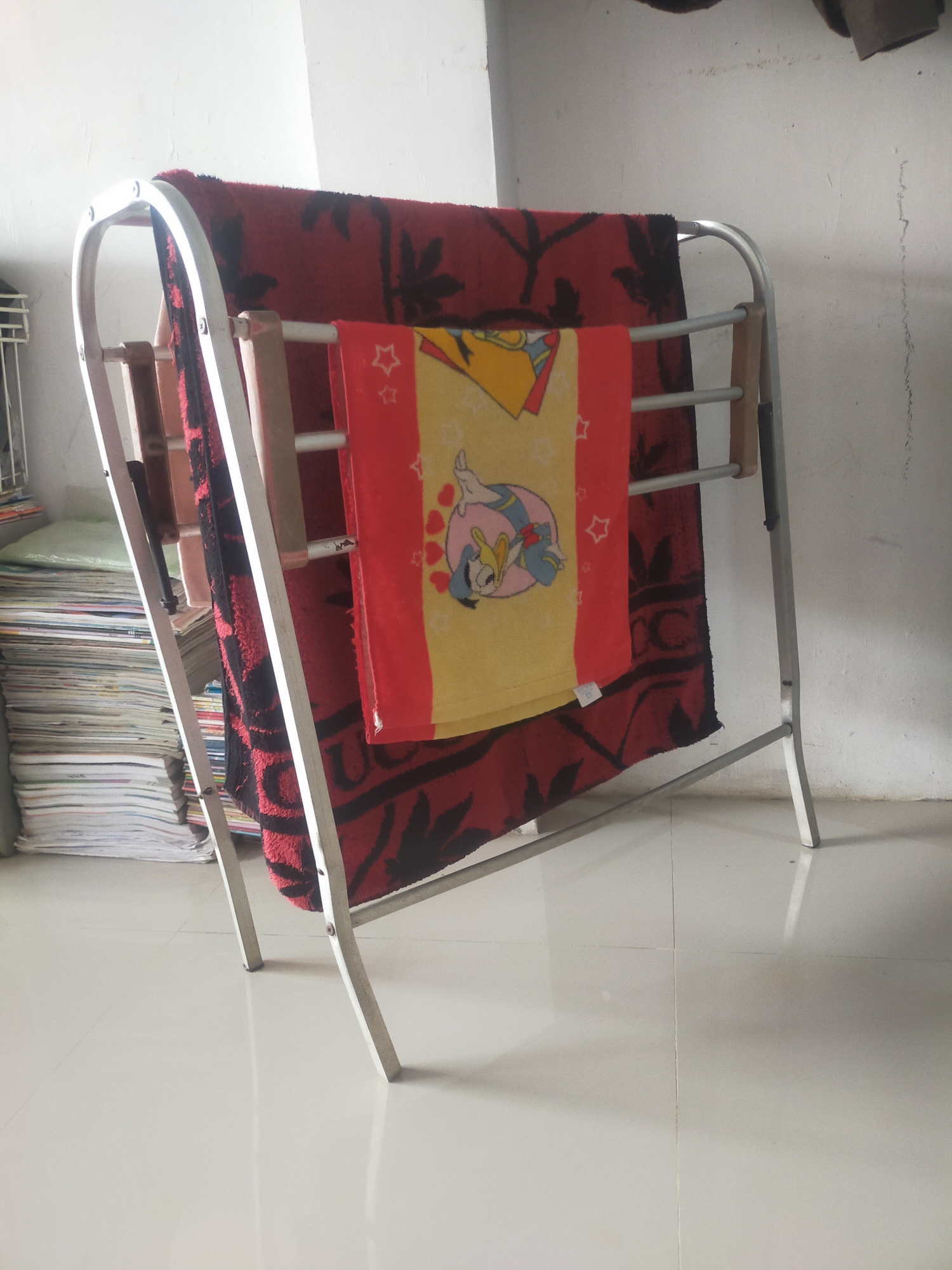 An indoor scene in a room in Tegalandong, Indonesia, features a silver metal drying rack holding two towels. A large, dark red towel with a black leaf pattern is draped over the top bars of the rack, extending down the back. A smaller, yellow and red towel with cartoon characters, possibly Donald Duck, is folded and placed on the front bars of the rack. To the left of the drying rack, a tall stack of papers or books is visible. The floor appears to be tiled in a light color, reflecting light. The background consists of a white wall with some faint vertical lines.