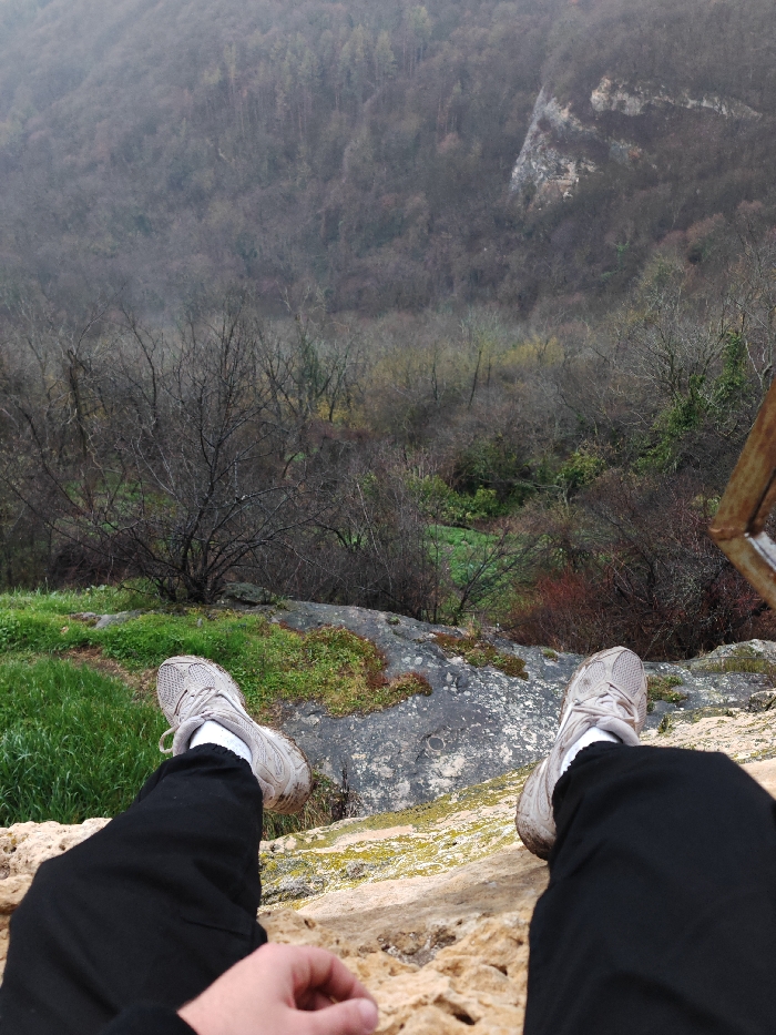 The image presents a first-person, downward view from a high vantage point on the Shumen Plateau, near Shumen, Bulgaria. In the immediate foreground, the legs and feet of an individual are visible, clad in black pants, white socks, and soiled light grey athletic shoes. The feet extend over the edge of a textured, light brown rock formation. A hand rests on this rock in the lower-left. Below the individual, a steep slope descends into a valley, characterized by patches of dark grey rock, bright green moss, and sparse, leafless deciduous trees and shrubs. The distant background reveals densely forested hills under an overcast sky, with a prominent grey rock face visible on the right slope. A fragment of a rusty, reddish-brown metallic structure, possibly a railing, is partially visible on the far right edge of the frame.