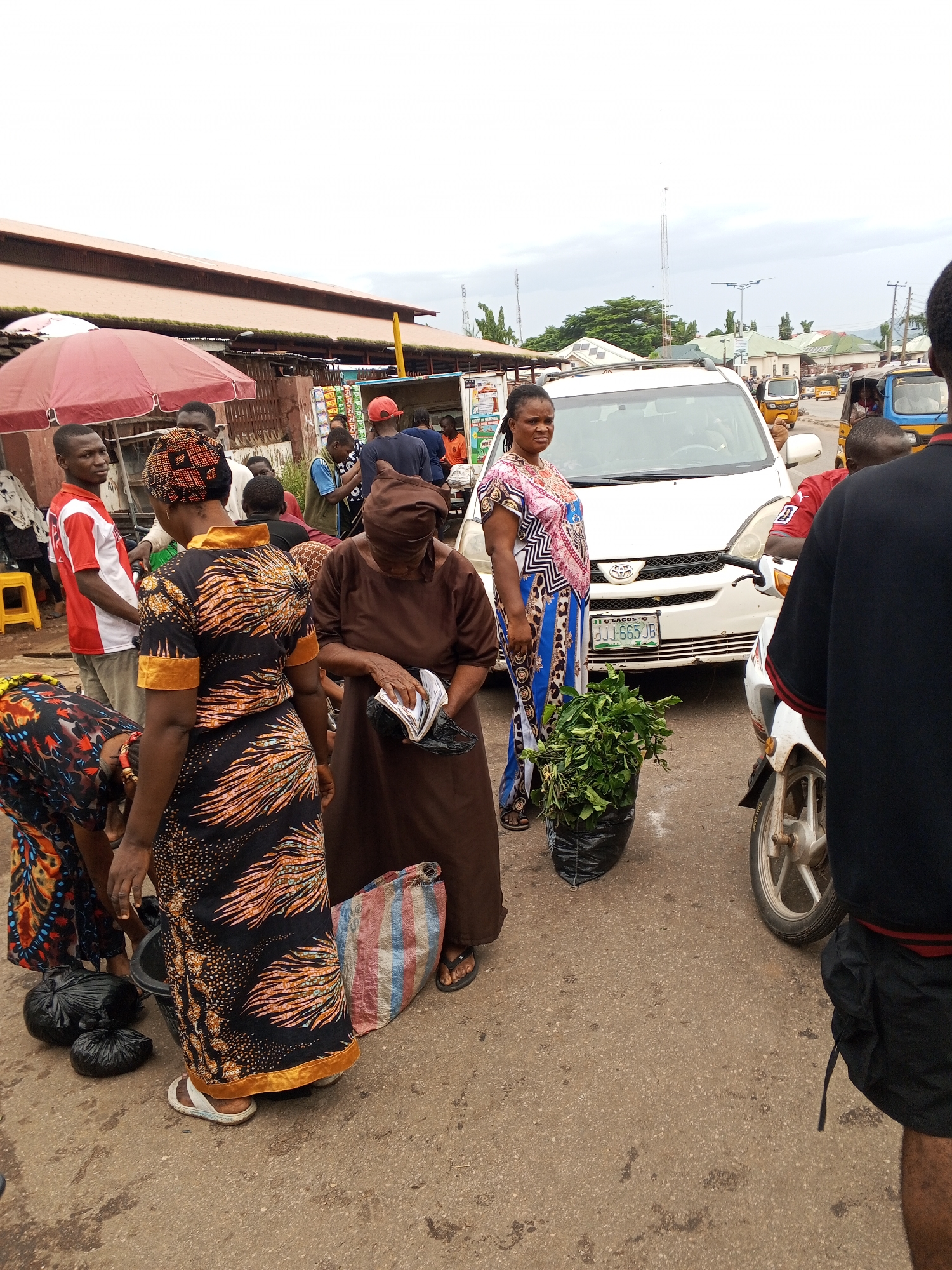 The image depicts a street scene in Lokoja, Nigeria, likely a market area. In the foreground, several people are gathered around a white Toyota minivan, which has the license plate "LAGOS DJJ 665 JB". To the left, a man in a red and white shirt stands near a red umbrella. A woman in a black and orange patterned dress is bending down, while another woman in a brown headscarf and brown dress is holding some papers and a black bag. Beside her, a large black plastic bag filled with green leafy vegetables is placed on the ground. To the right, a portion of a white motorcycle is visible, with its wheel in focus. In the background, more people, stalls, and vehicles, including yellow and blue tricycles, are present, suggesting a busy urban environment. The architecture includes a long building with a reddish-brown roof and some smaller structures with green roofs. The sky is overcast, indicating a cloudy day. The ground is paved with asphalt.